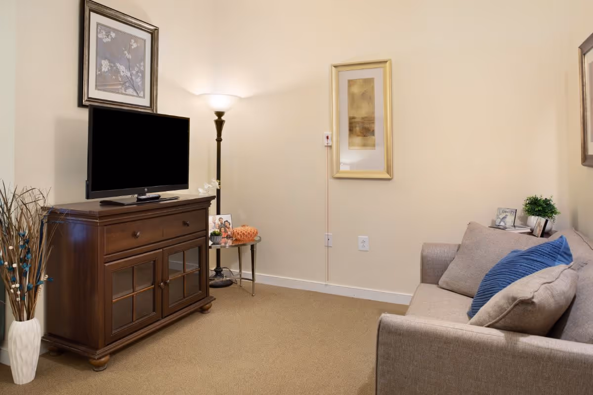 A cozy living room with a beige sofa adorned with blue and beige pillows, a wooden TV stand with a flat-screen television, a floor lamp, a small round side table with decorative items, and framed artwork on the walls.