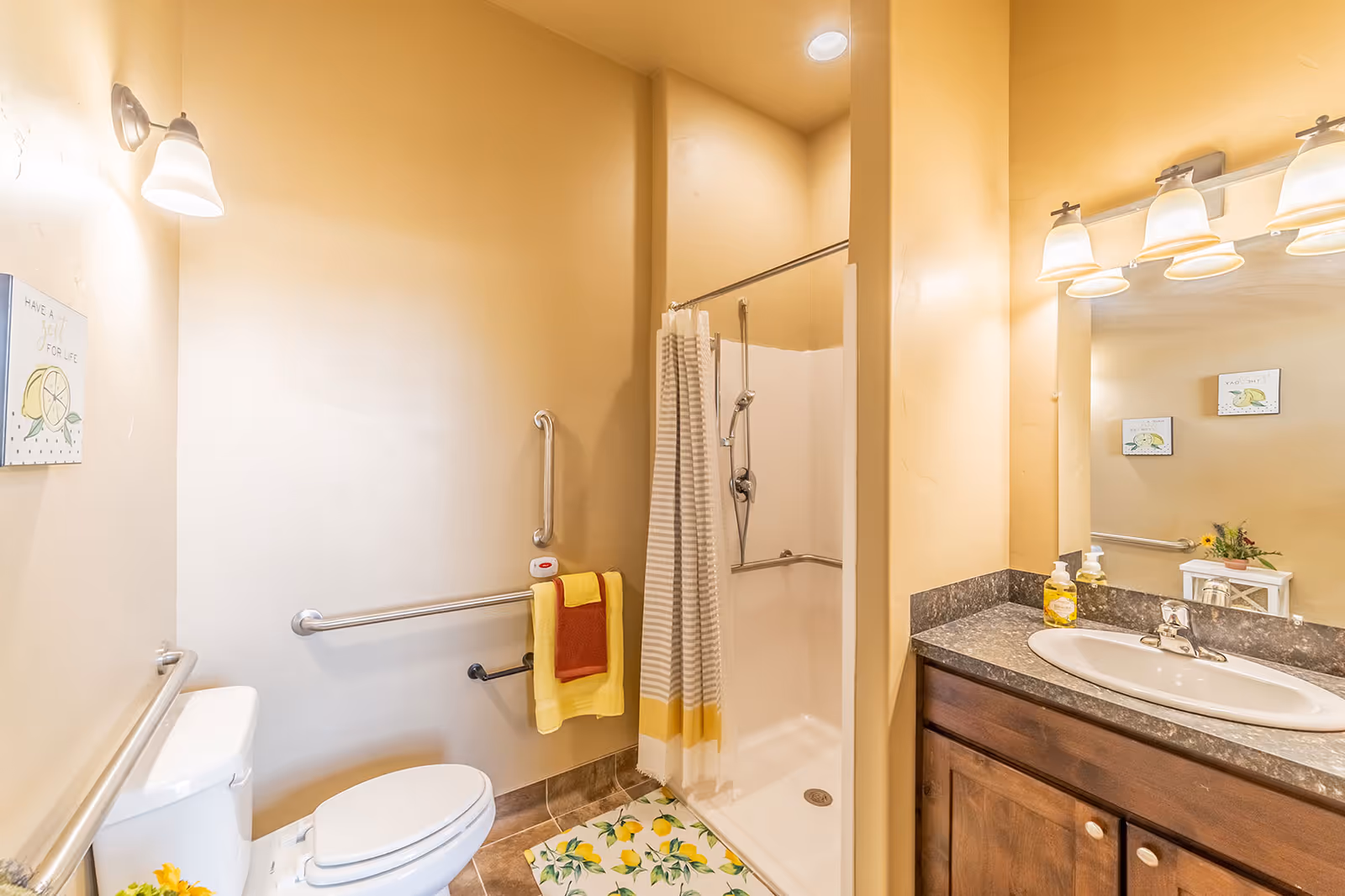 A well-lit bathroom with beige walls featuring a white toilet, a walk-in shower with a striped shower curtain, a sink with a dark countertop and wooden cabinet, and safety grab bars installed near the toilet and inside the shower. There are yellow and red towels hanging on the grab bars, a lemon-themed floor mat, and decorative lemon-themed wall art.