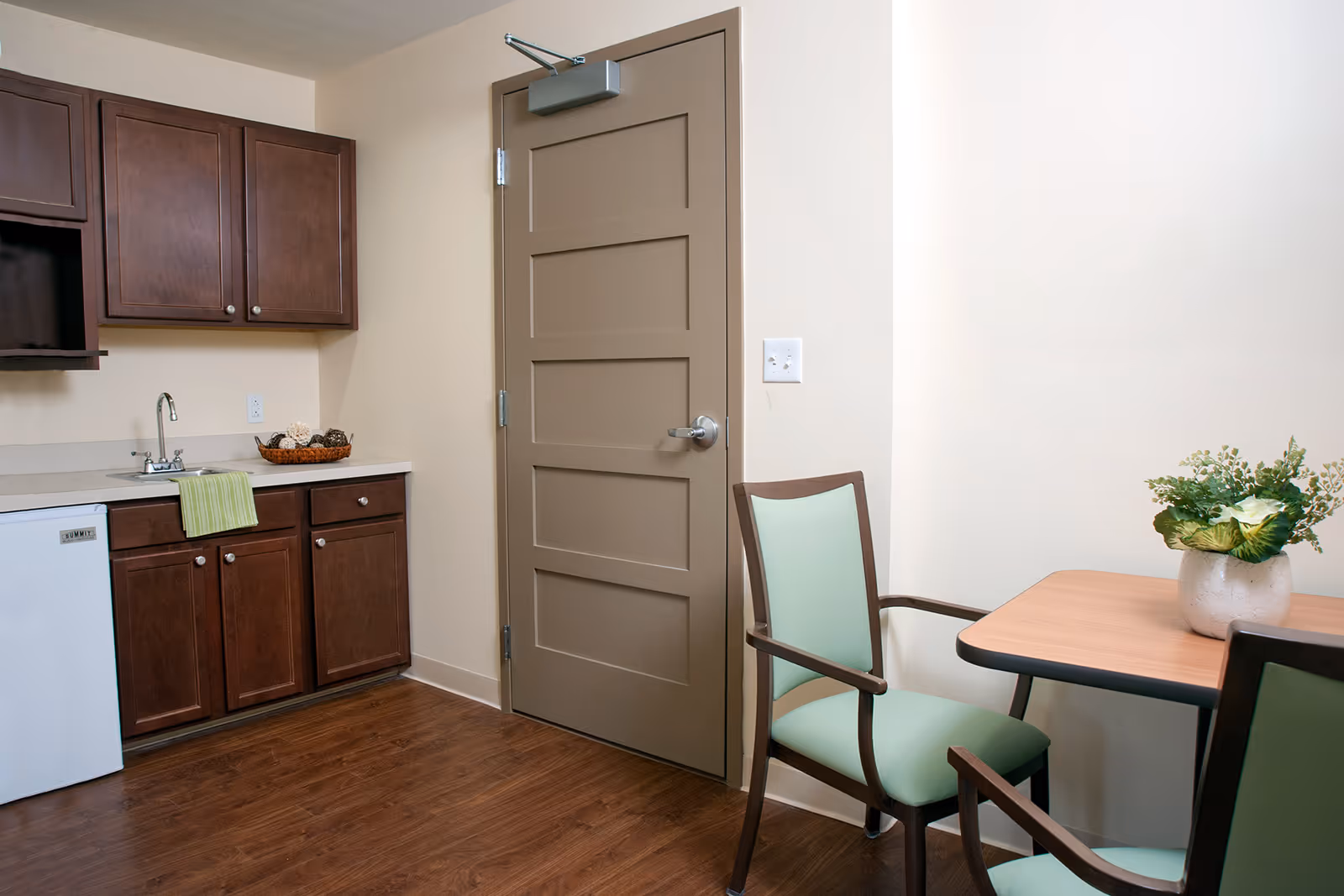 A small kitchenette area with dark wood cabinets, a white mini refrigerator, a sink with a green towel hanging over the edge, and a basket of decorative items on the counter. To the right, there is a brown door and a small wooden table with two chairs that have light green cushions. A vase with green and white flowers is on the table. The floor is wood and the walls are light beige.