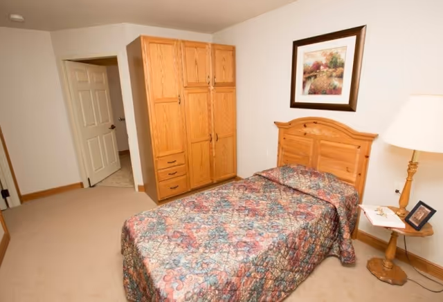 A bedroom with a single wooden bed covered with a multicolored floral quilt. Next to the bed is a wooden nightstand with a lamp, a small framed photo, and some papers. On the wall above the bed is a framed painting of a scenic landscape. Across from the bed is a large wooden wardrobe with multiple drawers and cabinets. The room has beige carpet and light-colored walls, with an open door leading to another room.