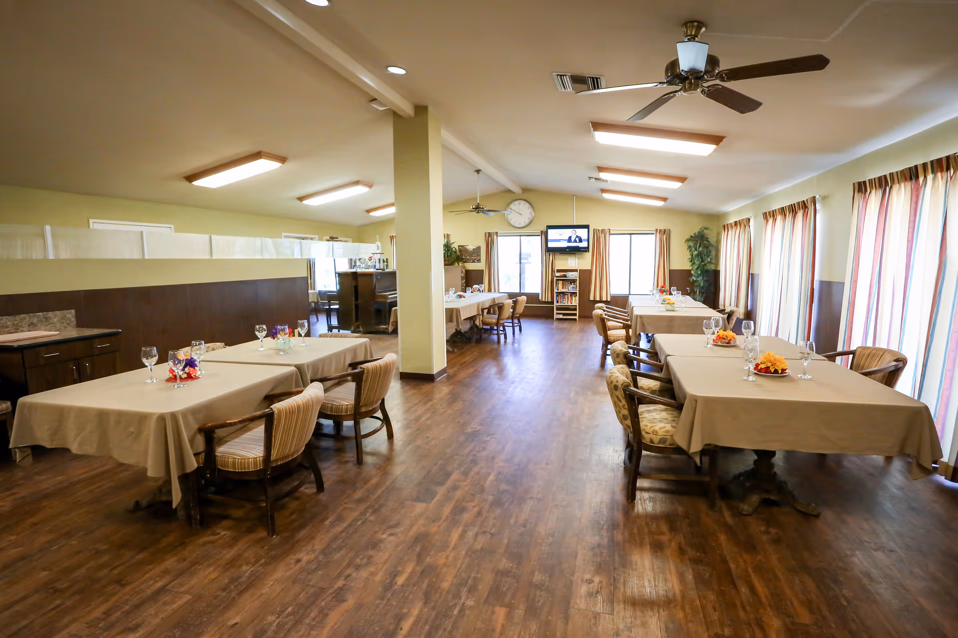 A spacious dining room with several tables covered with beige tablecloths, each set with glasses and small floral centerpieces. The room has wooden flooring, ceiling fans, and large windows with striped curtains allowing natural light to enter. A wall clock and a television mounted on the far wall are visible, along with a bookshelf and some plants.