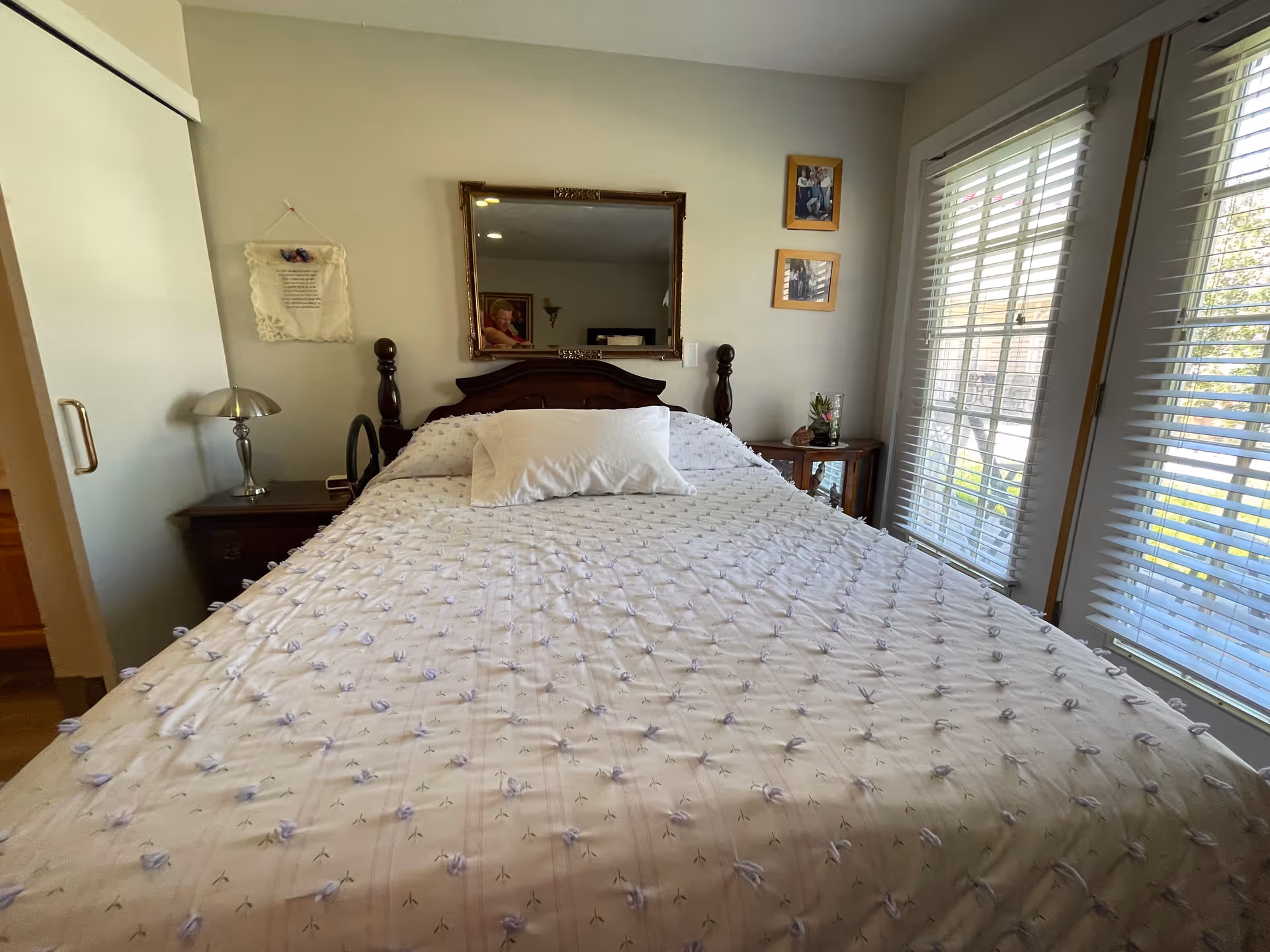 Sunlit bedroom with a made bed, wooden headboard, nightstands, and large windows with blinds.