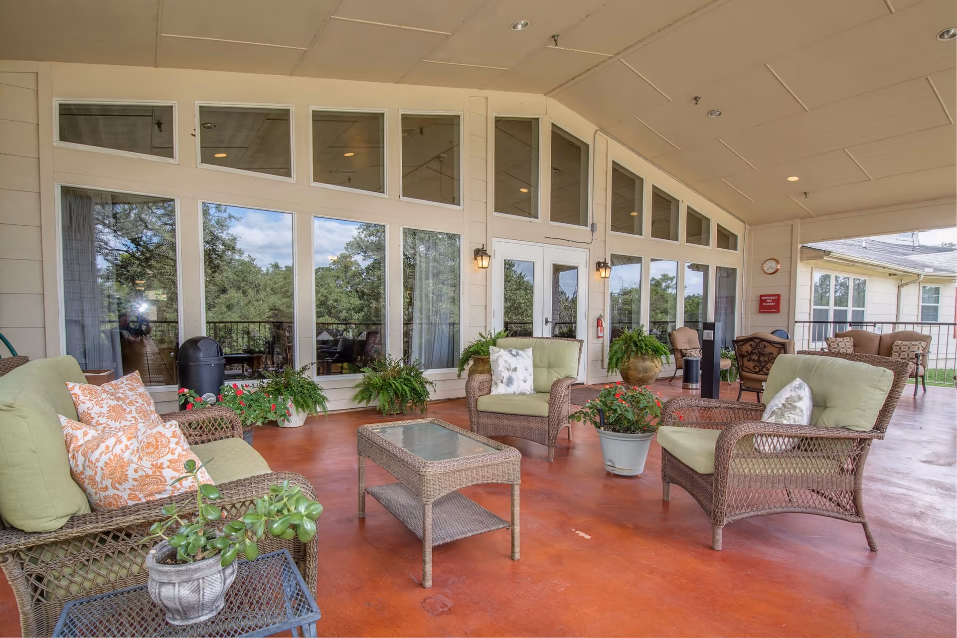Covered outdoor patio with wicker sofas and chairs, potted plants, and glass doors/windows along the building.