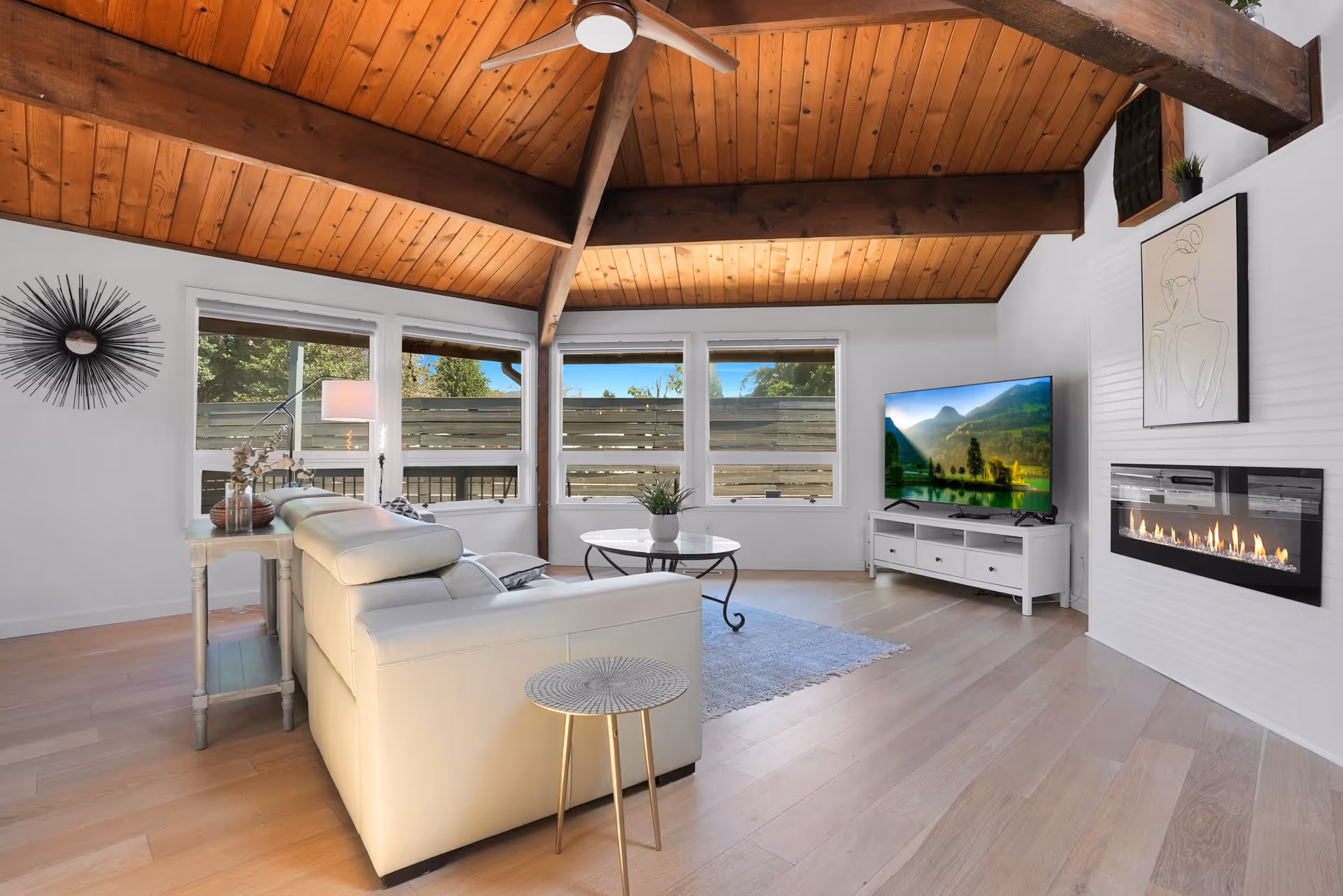 A cozy living room with a high wooden ceiling featuring exposed beams and a ceiling fan. The room has light wood flooring and white walls. There is a white leather sectional sofa facing a flat-screen TV on a white TV stand. A modern linear fireplace is built into the wall next to the TV. A round coffee table with a plant on it is placed in front of the sofa, and a small side table is beside the sofa. Large windows let in natural light and show a fenced outdoor area with greenery.