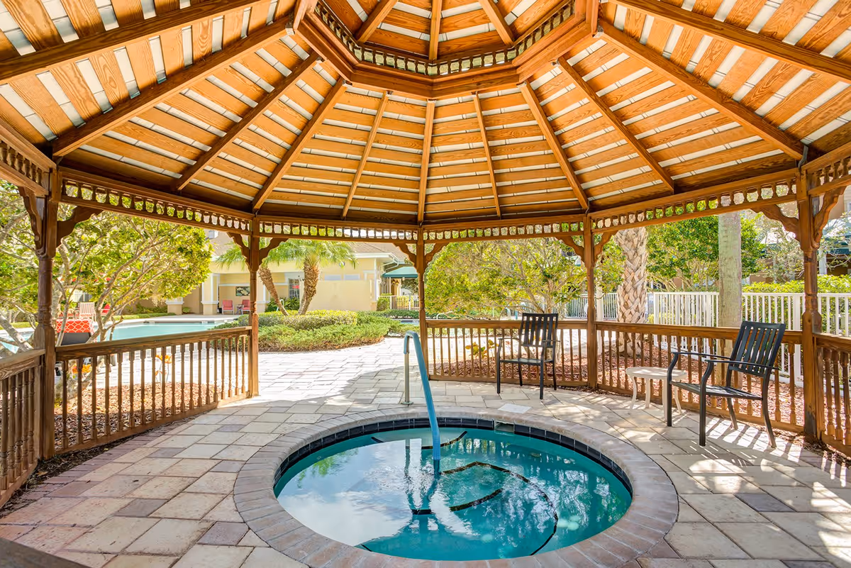 A wooden gazebo with a tiled roof covering a circular hot tub surrounded by a paved patio area. There are two black chairs and a small white table inside the gazebo. Outside the gazebo, there are trees, bushes, and a building in the background.
