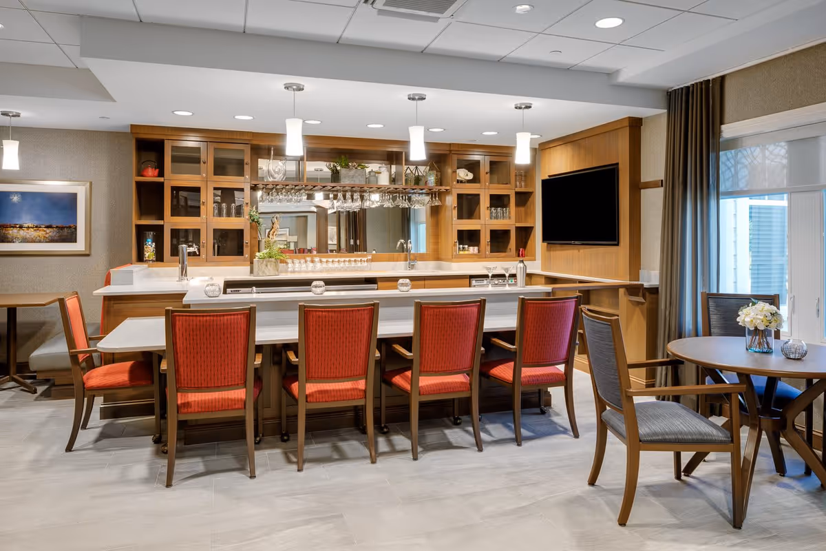A modern assisted living facility common area featuring a bar with six red cushioned chairs, wooden cabinetry with glass doors, hanging wine glasses, and a mounted flat-screen TV. To the right, there is a round table with two gray cushioned chairs and a vase with white flowers. The room has large windows with curtains allowing natural light to enter.