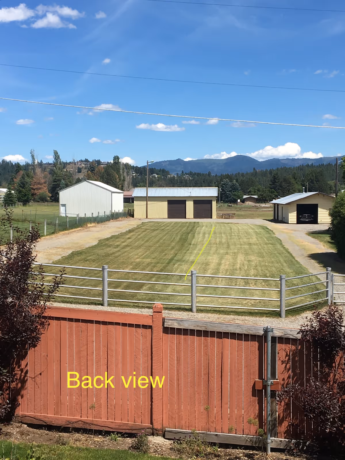 View of a fenced backyard with a red wooden gate in the foreground, a large grassy area, and three detached garage buildings in the distance under a blue sky with scattered clouds and mountains in the background.
