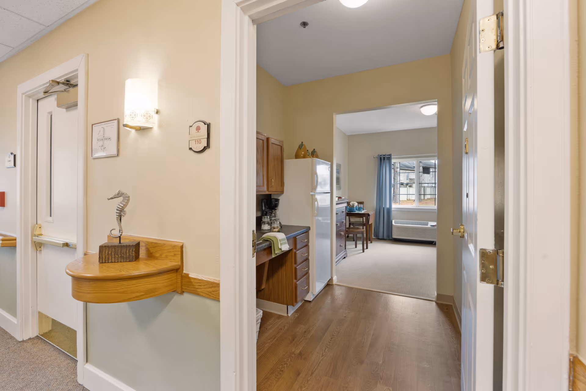 View through an open doorway into a senior living apartment at Discovery Commons Virginia Beach. The foreground shows a hallway with a small wooden shelf holding a decorative seahorse sculpture. Inside the apartment, there is a kitchenette with wooden cabinets, a white refrigerator, and a coffee maker. Beyond the kitchenette is a living area with a window covered by blue curtains, a small table with chairs, and a heating/cooling unit below the window. The walls are painted light beige, and the flooring transitions from wood in the kitchenette to carpet in the living area.