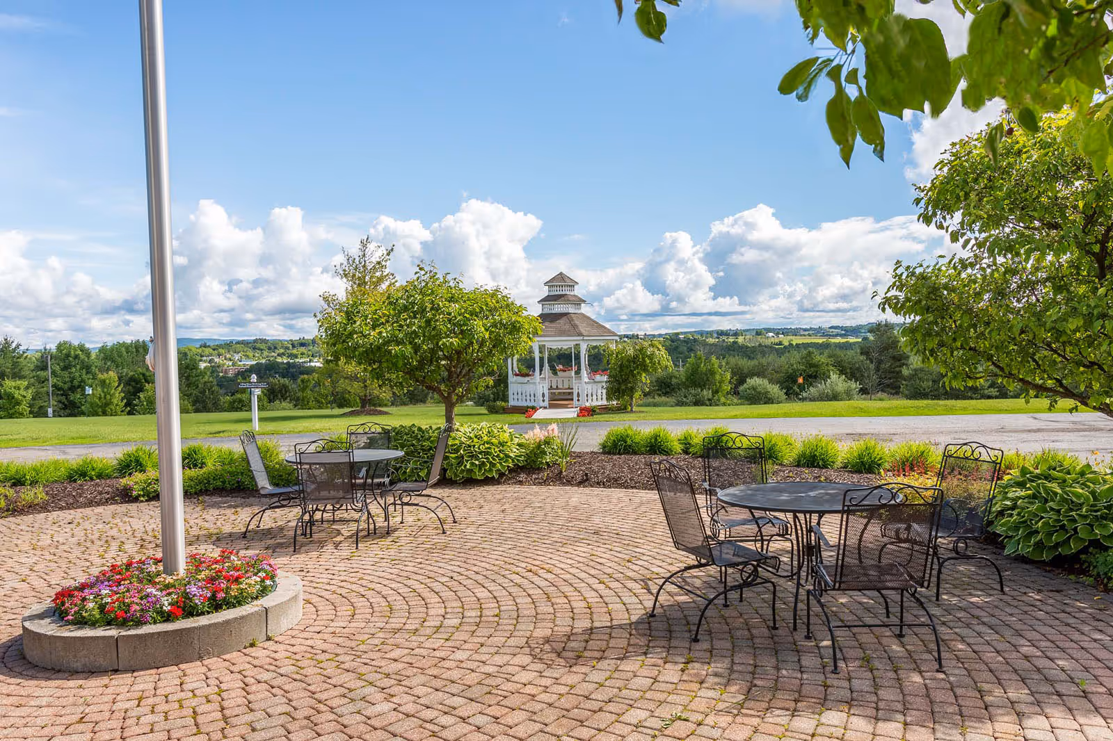 Outdoor patio area with round metal tables and chairs on a brick-paved surface, surrounded by green trees and plants. In the background, there is a white gazebo with a peaked roof, set against a scenic view of a green landscape and a partly cloudy blue sky.