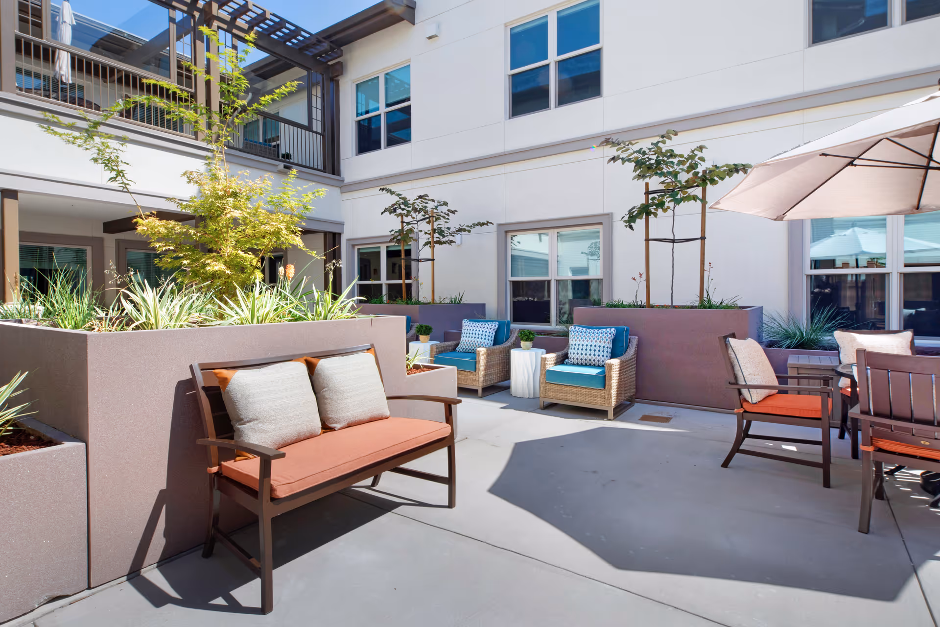 Outdoor courtyard area with seating including a bench with cushions, wicker chairs with blue cushions, and a table with chairs under a large umbrella. There are raised planters with small trees and plants, surrounded by a two-story building with windows.