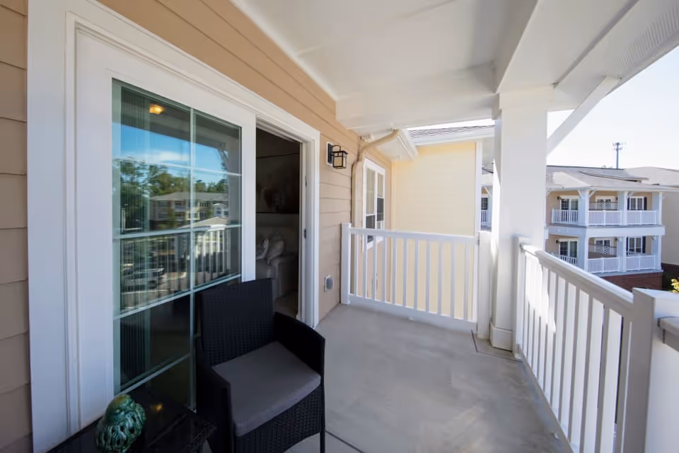 A covered balcony with white railings and a beige exterior wall. There is a black wicker chair with a gray cushion and a small black table with a decorative item on it. The balcony overlooks other residential buildings with similar balconies.