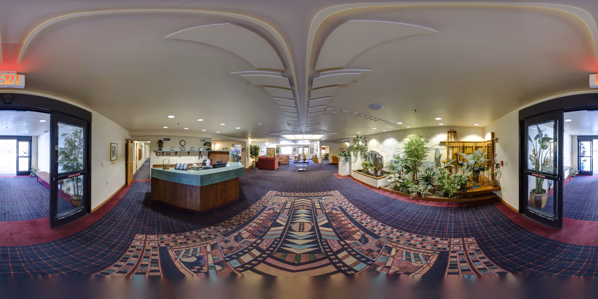 Wide panoramic view of a senior living facility lobby with a reception desk in the center, decorative plants on the right, seating areas in the background, and double glass doors on both sides. The floor is carpeted with a patterned design, and the ceiling has recessed lighting.