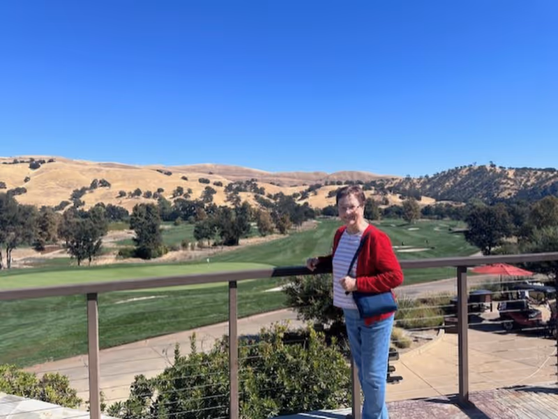 An elderly woman wearing a red jacket and blue jeans stands on a balcony overlooking a golf course with green grass, trees, and rolling hills under a clear blue sky.