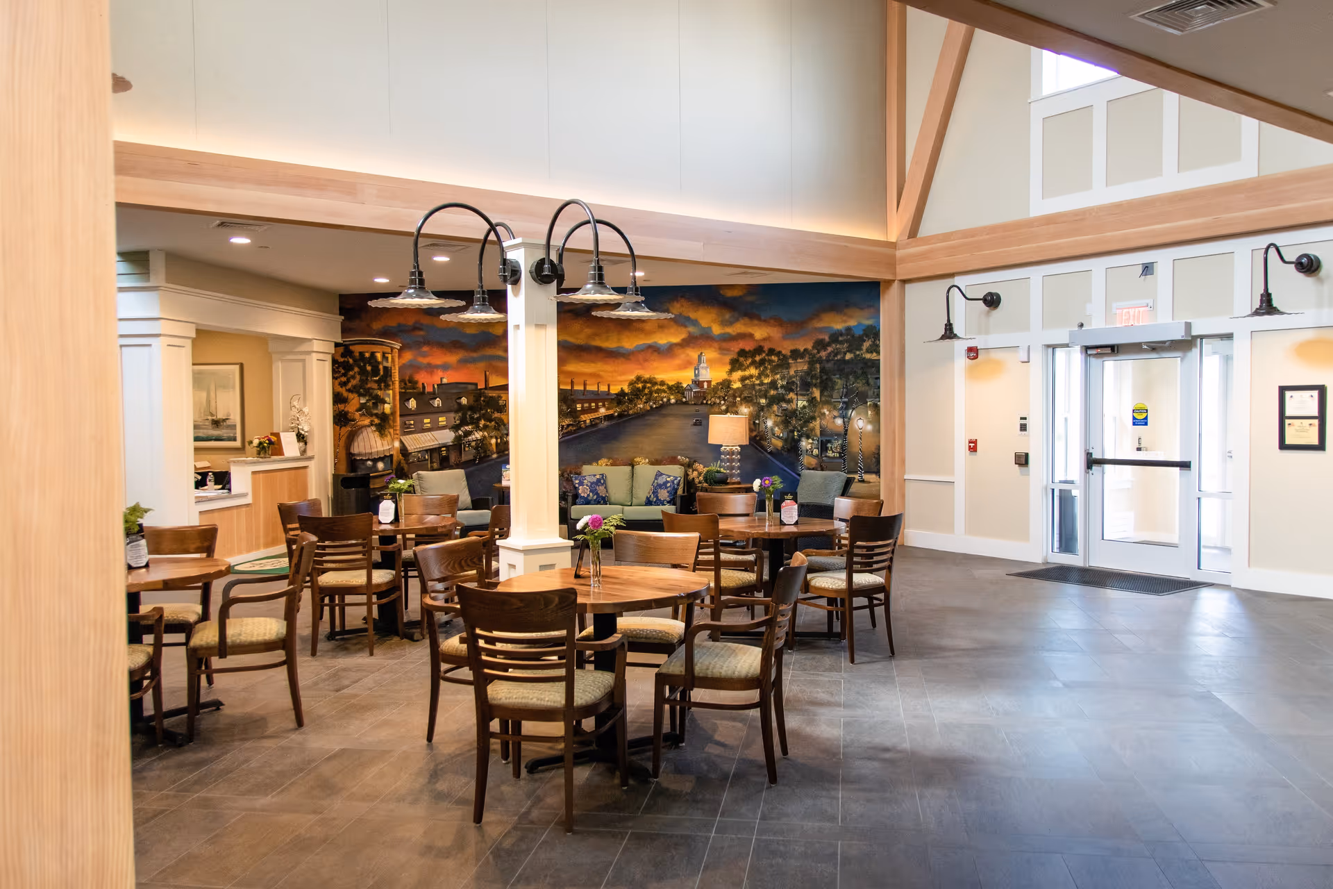 Interior view of a senior living facility common area with several round wooden tables and chairs arranged on a tiled floor. A large mural depicting a sunset over a town street decorates the back wall. There is a seating area with sofas and a lamp beneath the mural. The space has high ceilings with wooden beams and multiple light fixtures. An entrance door with glass panels is visible on the right side.