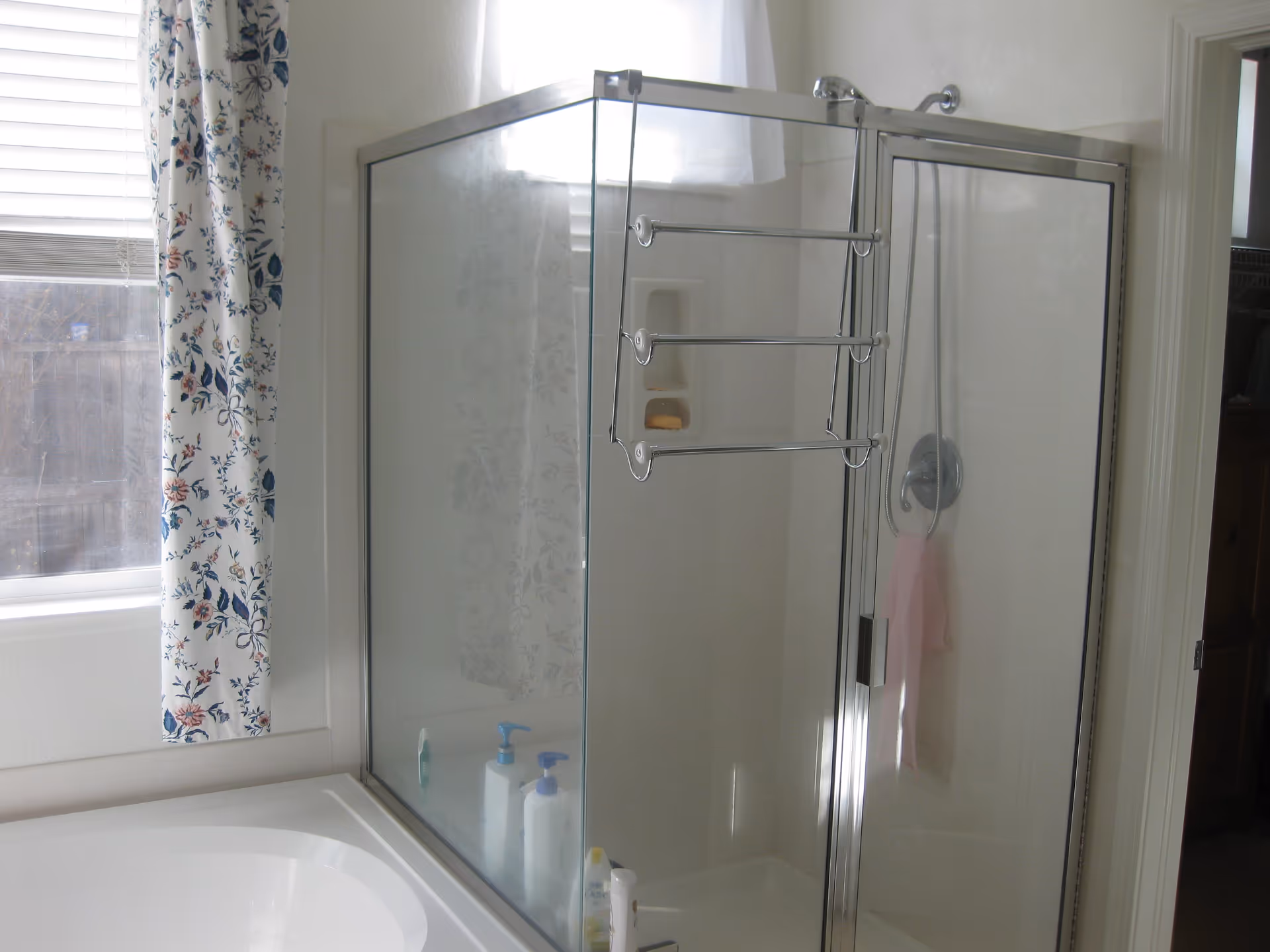 A bathroom with a glass-enclosed shower featuring a metal frame and a towel rack attached to the door. Next to the shower is a white bathtub. A window with floral-patterned curtains and blinds is visible on the left side, allowing natural light into the room.
