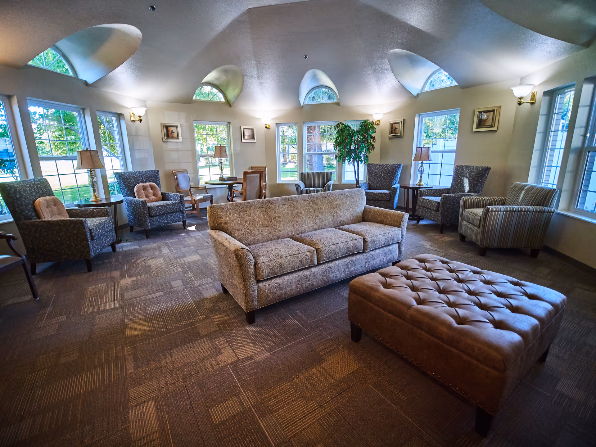A bright and spacious common area with multiple armchairs, a patterned sofa, and a large tufted ottoman. The room features large windows with views of greenery outside, arched windows near the ceiling, wall lamps, framed artwork, and a potted plant in the corner.