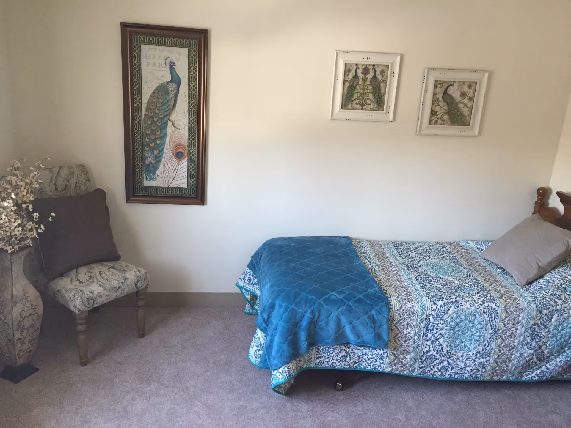 A bedroom with a single bed covered in a patterned blue and white bedspread and a blue blanket. There is a gray pillow on the bed. To the left, there is a decorative chair with a dark cushion and a tall vase with white flowers. Three framed pictures of peacocks hang on the wall above the bed and chair.
