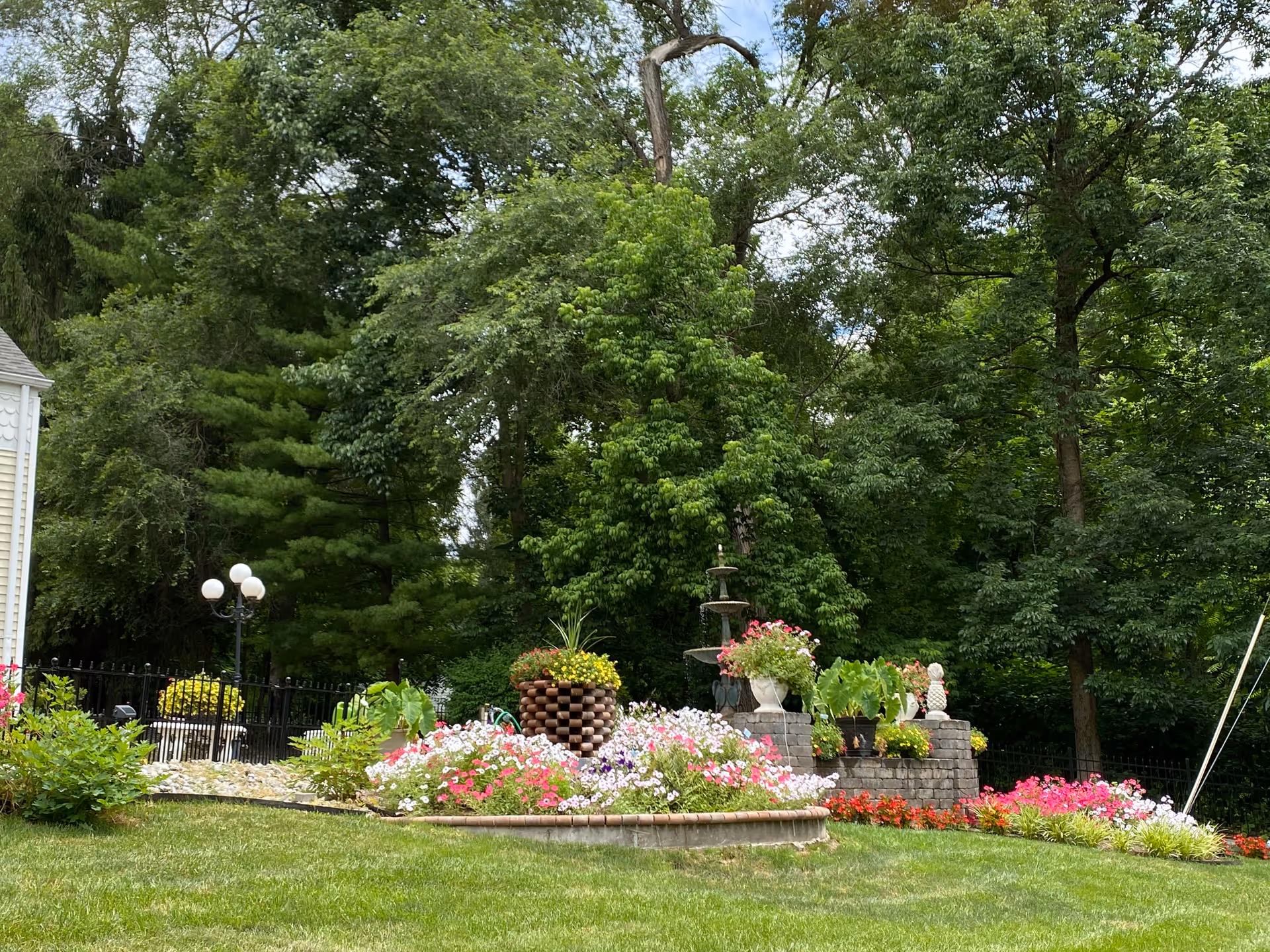 A landscaped garden area with a variety of colorful flowers and plants arranged around a tiered water fountain. The garden is surrounded by lush green trees and a well-maintained lawn. There is a black metal fence and a lamp post with three white globes visible in the background.