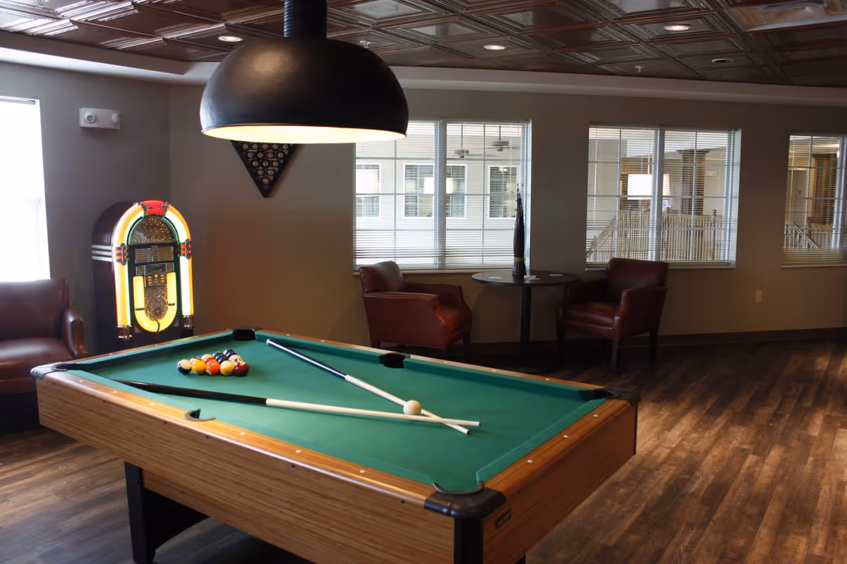 A recreational lounge with a pool table, hanging light, a jukebox, and leather chairs by windows.