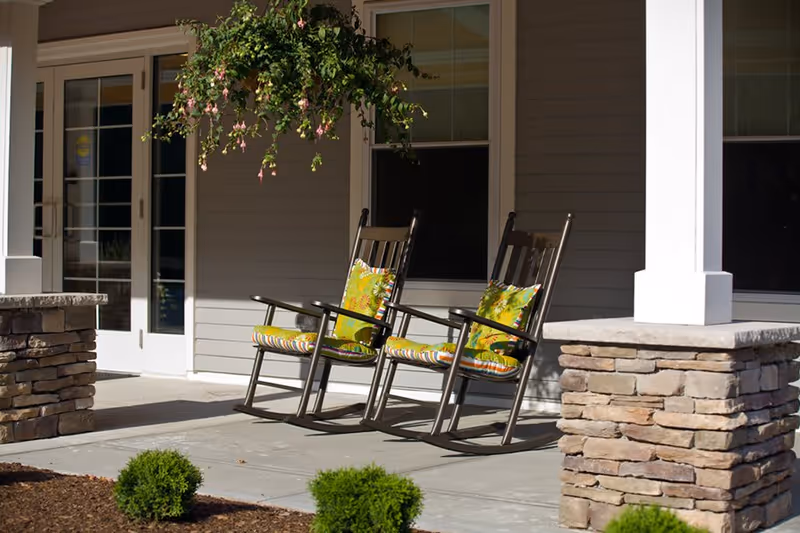 Two wooden rocking chairs with colorful cushions on a porch with stone pillars and a hanging plant, outside a building with gray siding and white trim.