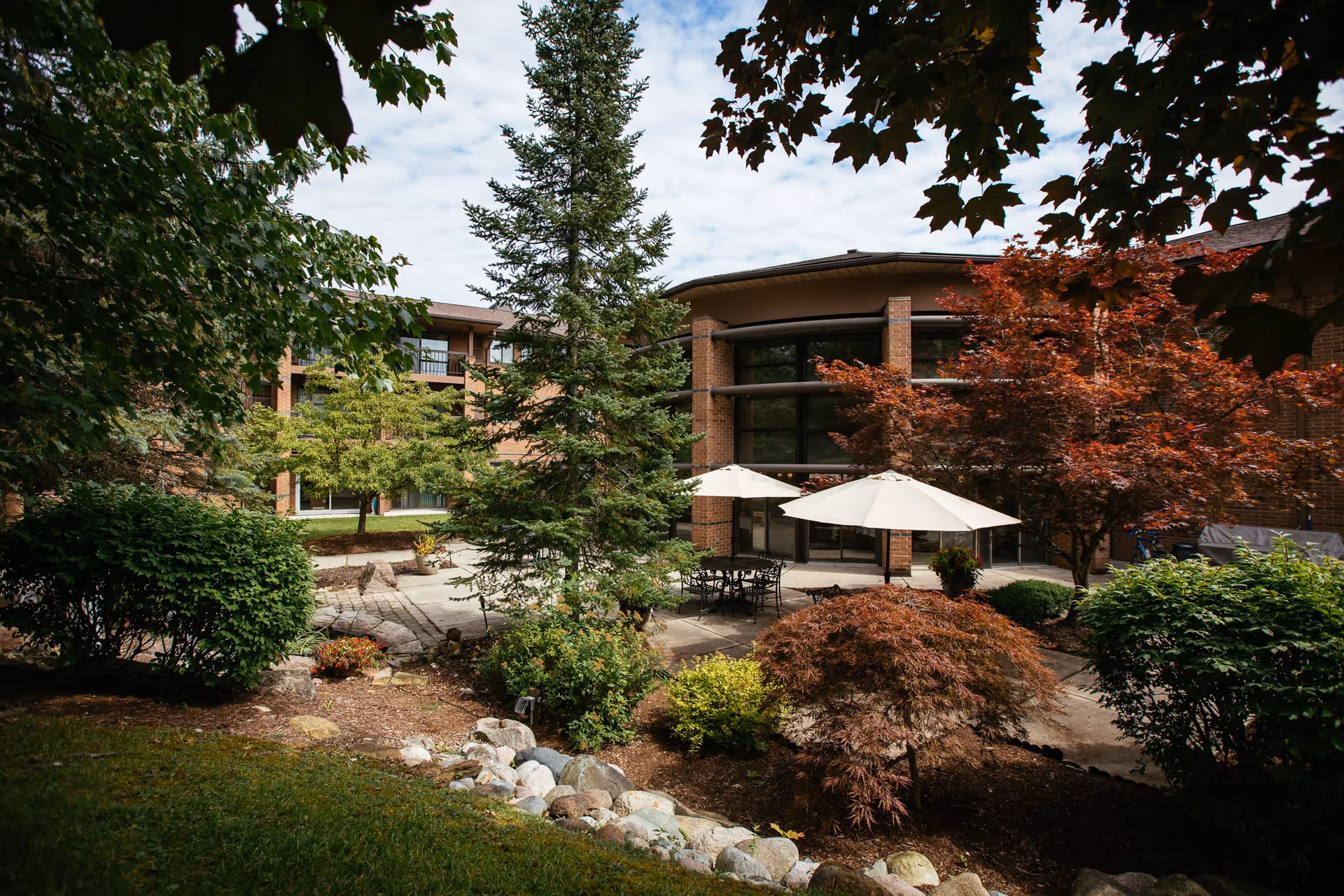 Outdoor courtyard area of a senior living facility with various trees and shrubs, a paved patio with tables and chairs under white umbrellas, and a multi-story brick building in the background under a partly cloudy sky.