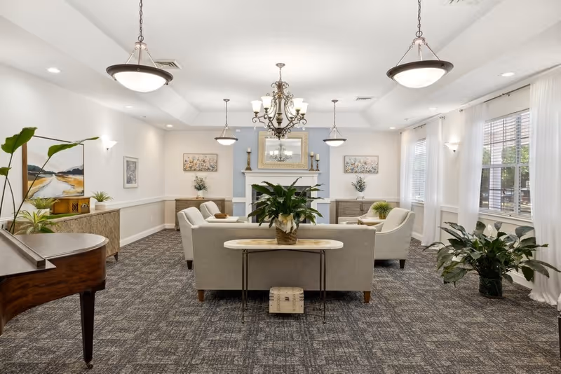 A bright and spacious living room area with a gray carpet and white walls. The room features a central seating arrangement with a sofa and armchairs around a fireplace with a large mirror above it. There are several plants, a grand piano on the left, and large windows with white curtains letting in natural light. Elegant chandeliers and pendant lights hang from the ceiling.