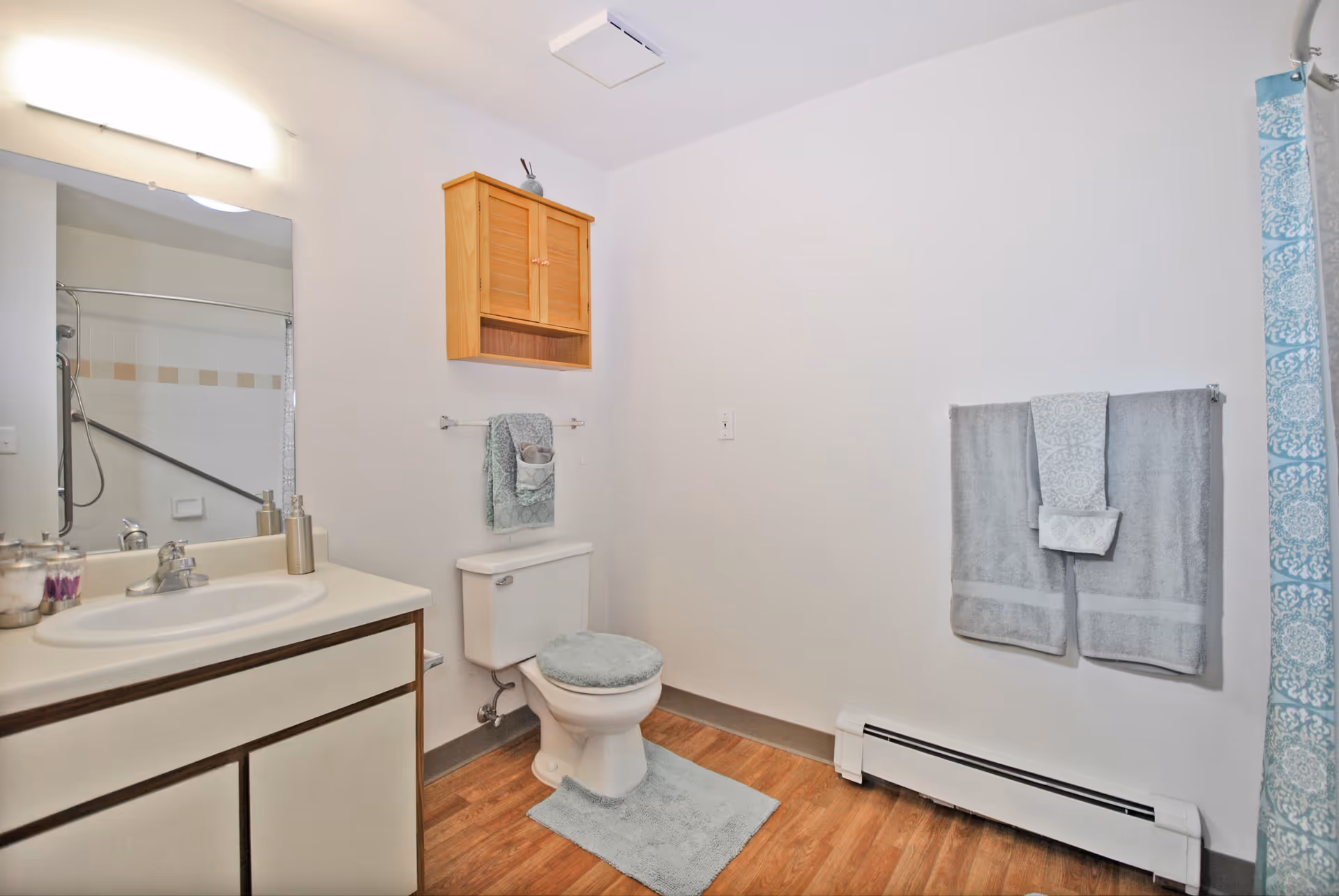 A clean bathroom with a white toilet covered with a light blue fuzzy seat cover and matching bath mat. Above the toilet is a wooden cabinet and a towel rack with a folded towel. To the left is a sink with a mirror above it and a soap dispenser on the counter. On the right wall, two gray towels are hanging on a towel rack next to a blue and white patterned shower curtain. The floor is wood-style and the walls are white.