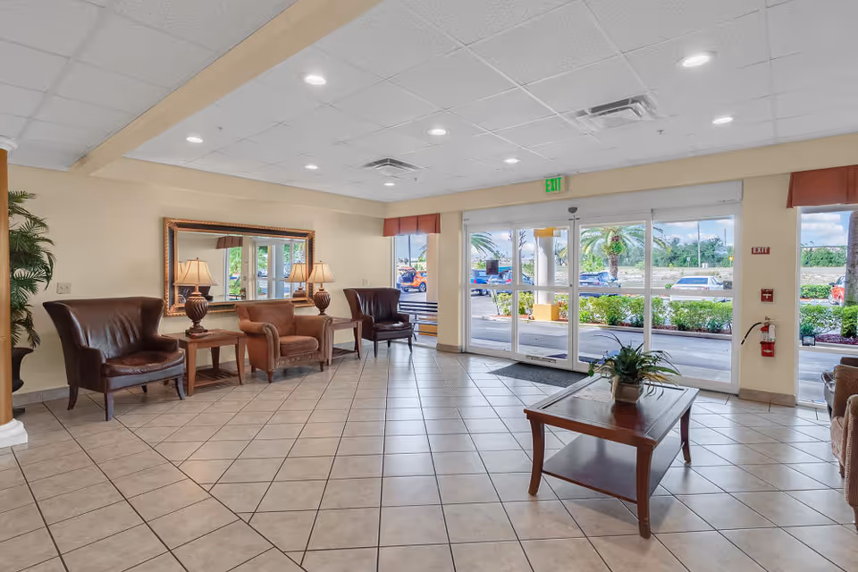 Spacious assisted living lobby with chairs, side tables and lamps, a coffee table, and large glass entrance doors showing the exterior landscaping.