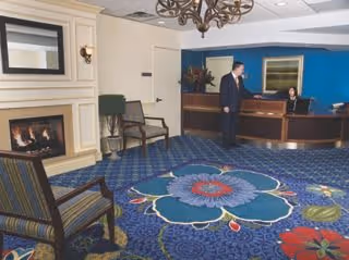 A cozy assisted living facility lobby with a blue floral carpet, a fireplace on the left, two chairs, a chandelier, and a reception desk where a man in a suit is talking to a receptionist.