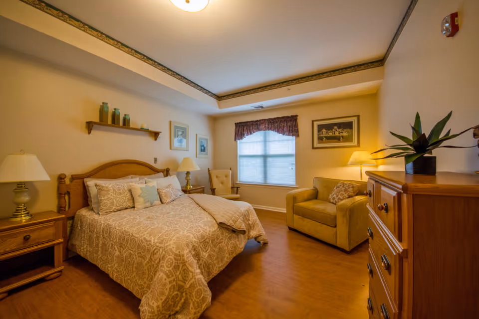 A cozy bedroom with a wooden bed frame, patterned beige bedspread, and multiple pillows. There are two bedside tables with lamps, a wooden dresser with a plant on top, a cushioned armchair, and a window with blinds and a valance. The walls are decorated with framed pictures and a small shelf holding decorative items.