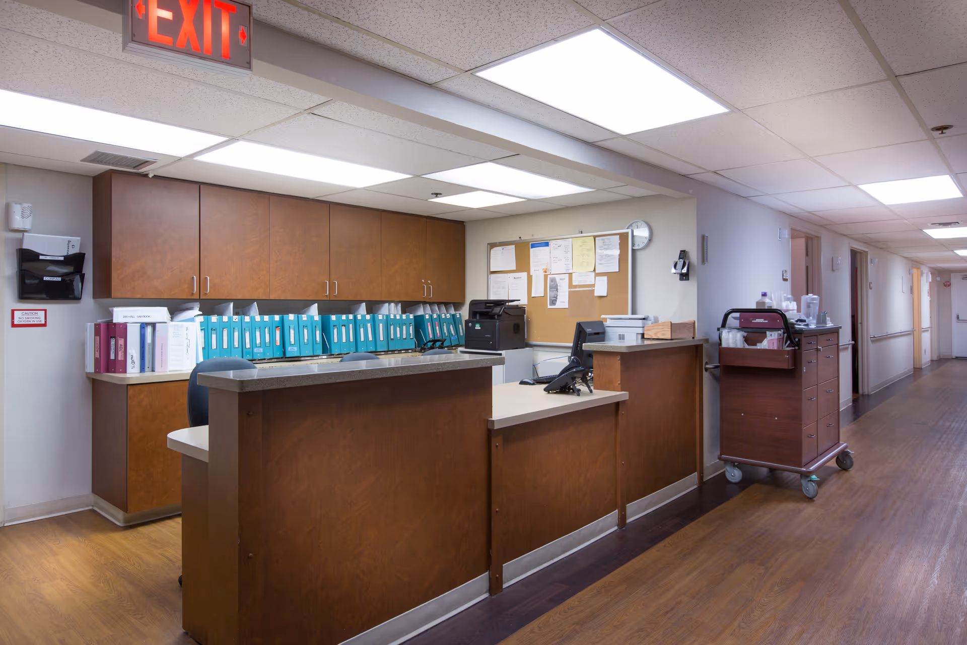 Reception desk area in a healthcare facility hallway with wooden cabinets, a bulletin board with papers, a computer, and a cart with medical supplies. The hallway extends to the right with doors along the wall and an illuminated exit sign on the ceiling.