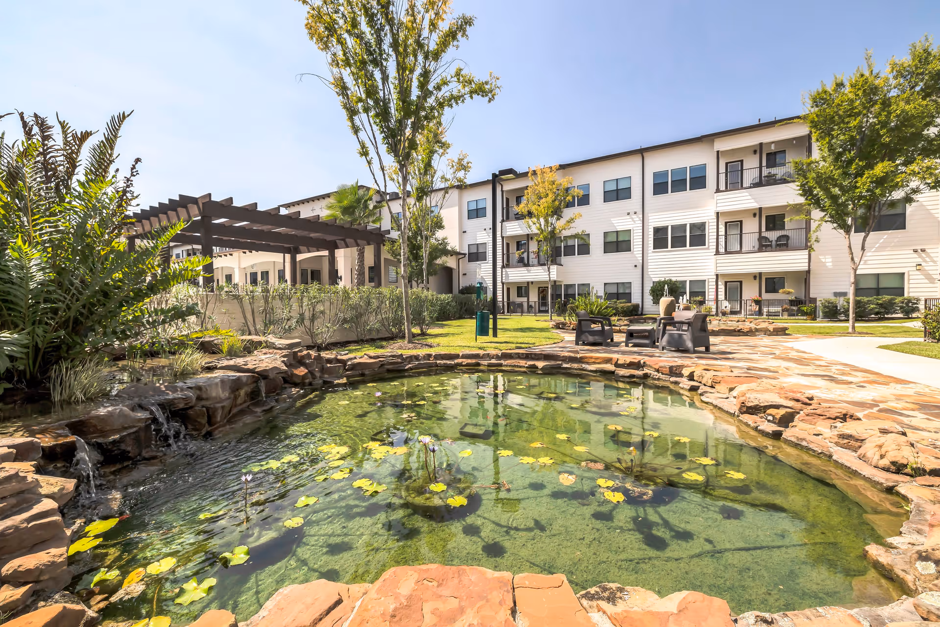 Outdoor courtyard featuring a decorative lily pond, seating area, pergola, and a three-story apartment building in the background.