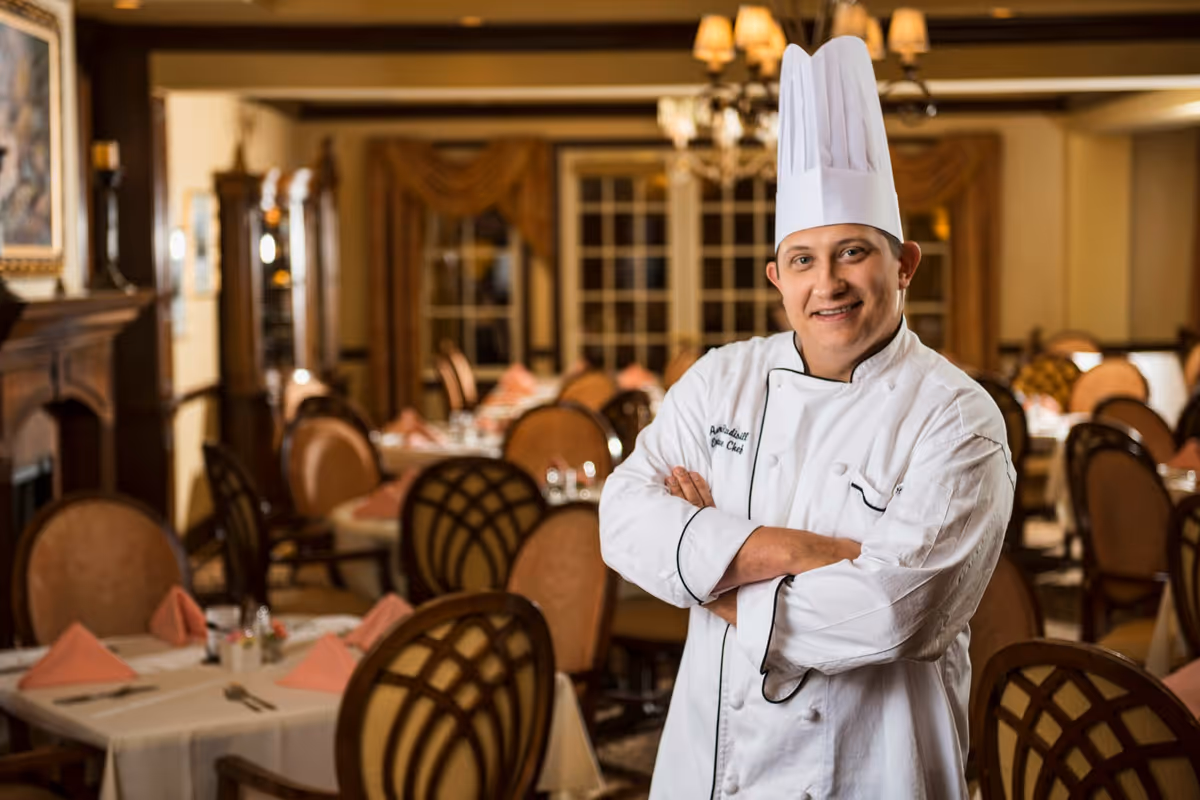 A chef wearing a white chef's coat and tall white chef hat stands with arms crossed in a warmly lit dining room with tables set with pink napkins and chairs with decorative backs.