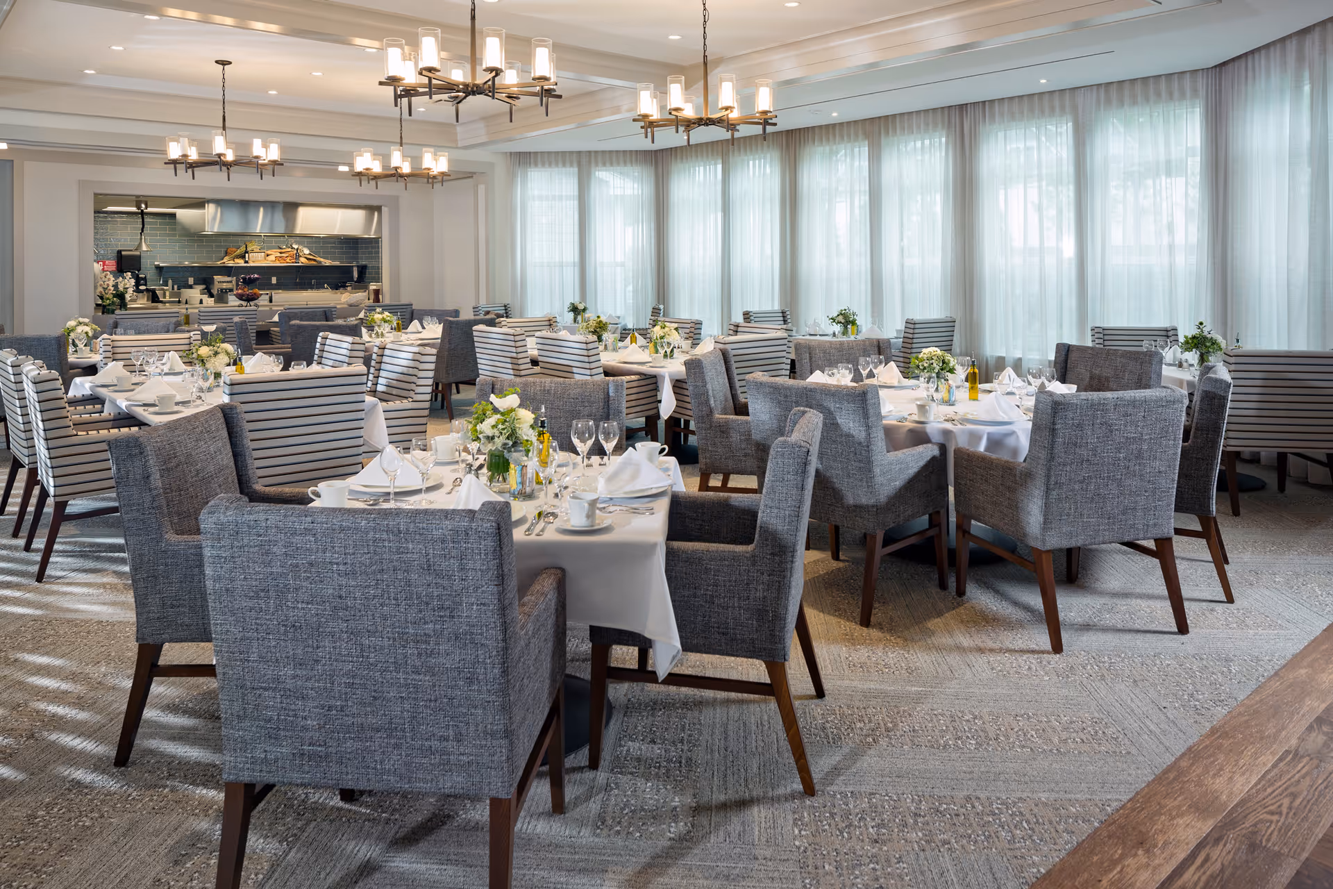 A bright and elegant dining room with multiple tables set for a meal. Each table is covered with a white tablecloth and decorated with floral centerpieces, glassware, and neatly arranged silverware. The chairs are upholstered in gray fabric, and large windows with sheer curtains allow natural light to fill the space. Modern chandeliers hang from the ceiling, and an open kitchen area is visible in the background.