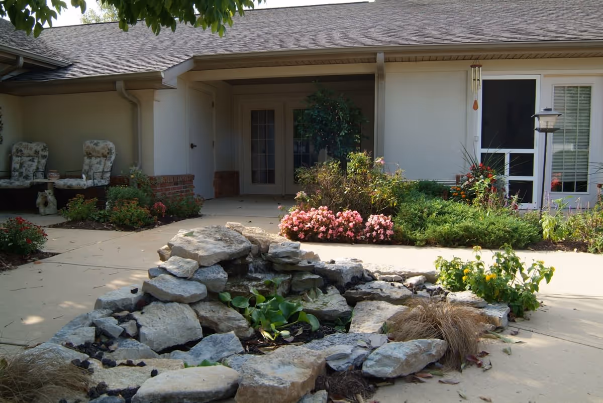 Courtyard entrance of a senior living building with a rock water feature, flower beds, and patio chairs.