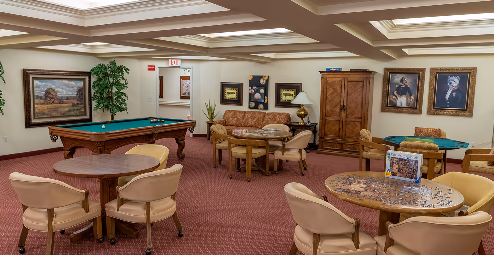 A recreational room with a pool table, several round tables with chairs, a sofa, framed artwork on the walls, and a puzzle in progress on one of the tables. The room has a coffered ceiling and carpeted floor.