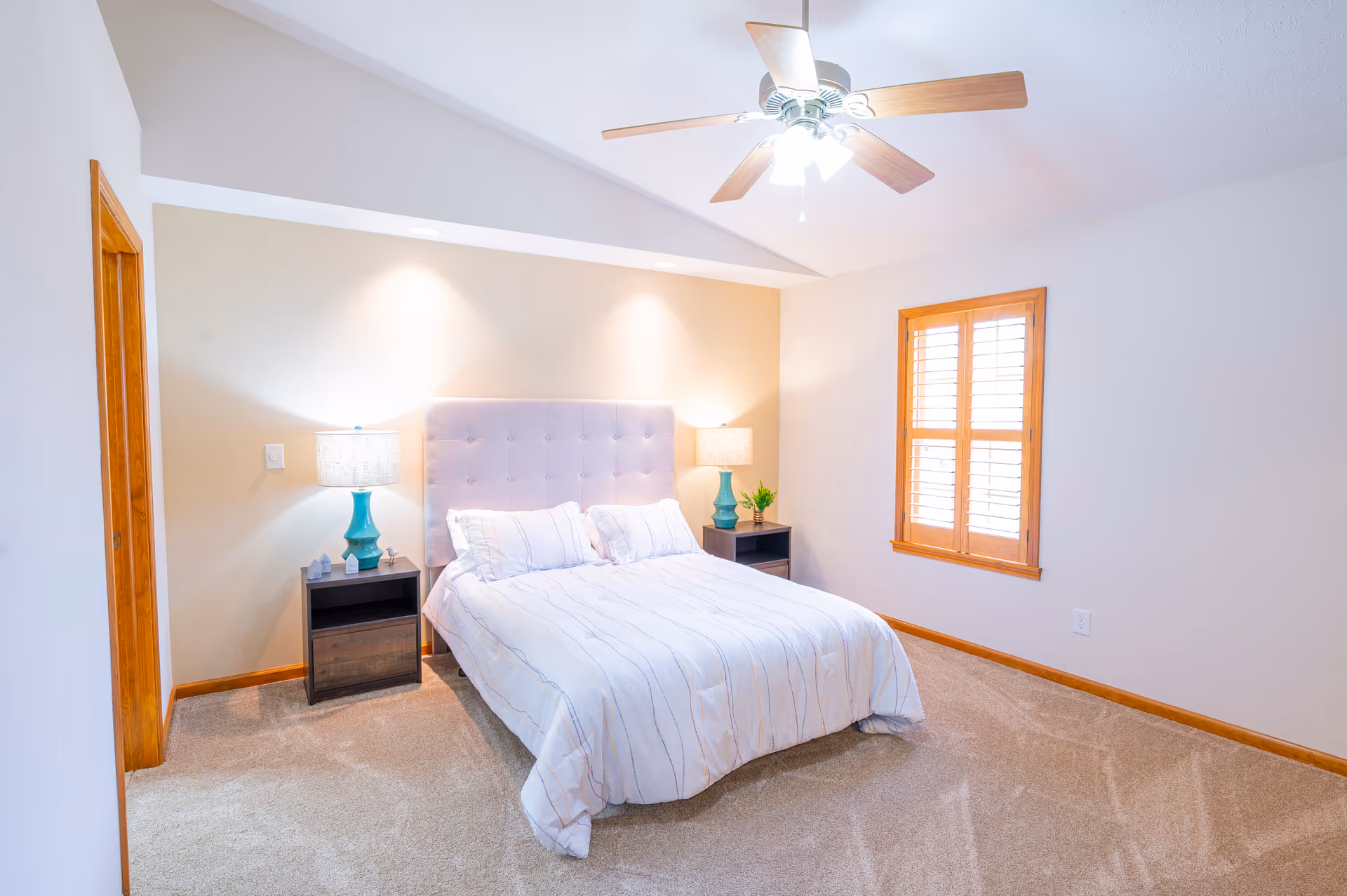 A bright bedroom with a queen-sized bed featuring a tufted headboard and white bedding with subtle stripes. There are two dark wooden nightstands on either side of the bed, each with a turquoise lamp and one with a small plant. The room has beige carpet, a ceiling fan with lights, and a window with wooden shutters.