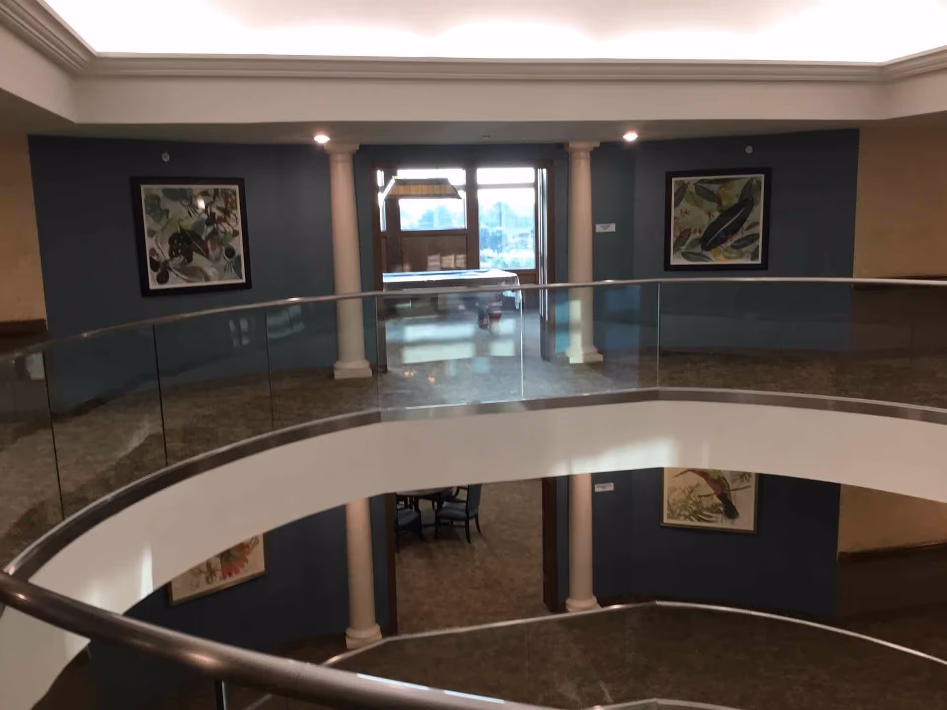 Interior view of a two-story area in a retirement community with a curved glass railing on the upper floor, beige carpet, blue walls, white columns, and framed artwork featuring nature themes. There is a doorway leading to a room with a table and chairs, and natural light coming through windows in the background.