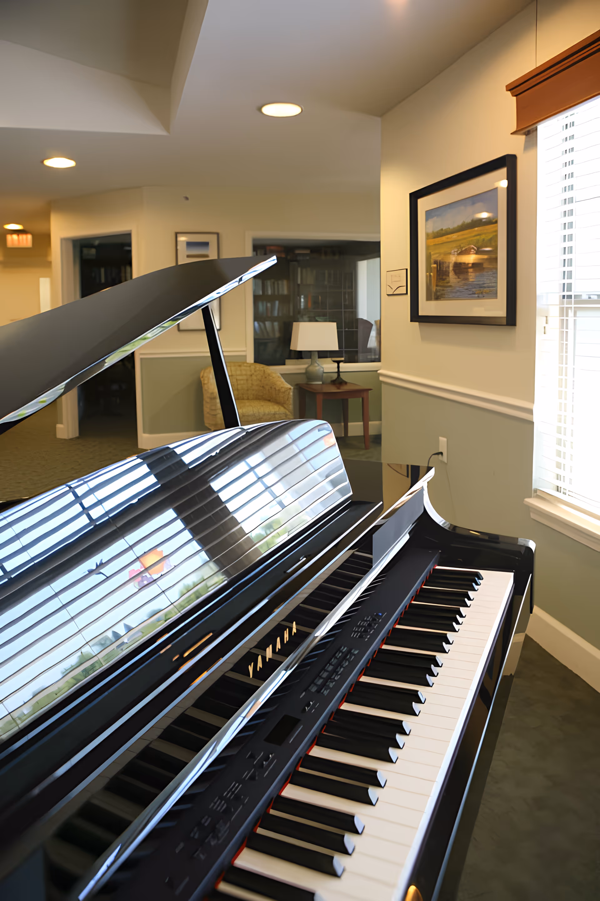 Black grand piano in a bright communal living area with framed art, an armchair, side table, and window blinds.