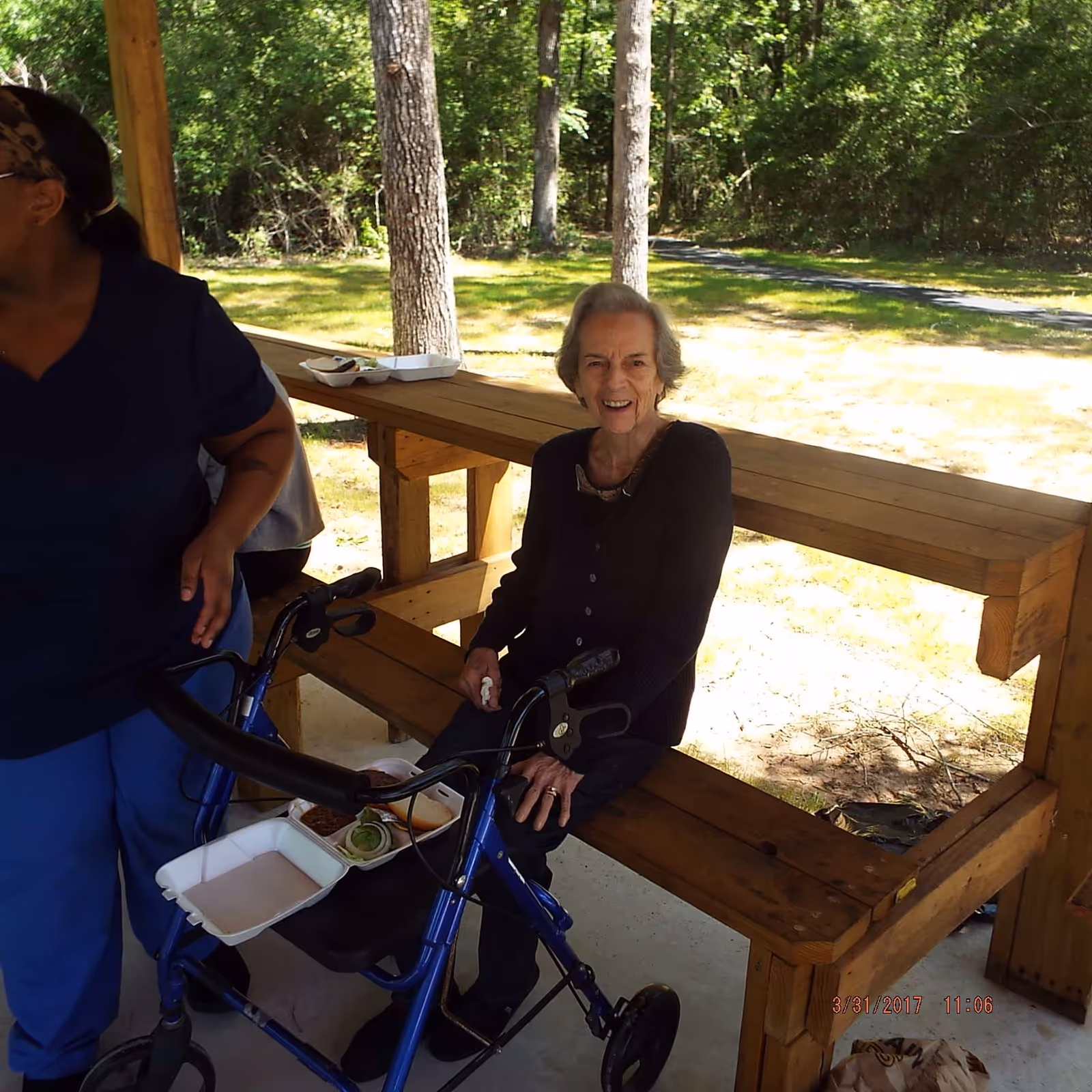 An elderly woman sitting on a wooden bench under a covered outdoor area, smiling and holding a tissue. She has a blue walker in front of her with a takeout food container resting on it. Another person wearing blue scrubs is partially visible standing nearby. Trees and a grassy area are visible in the background.