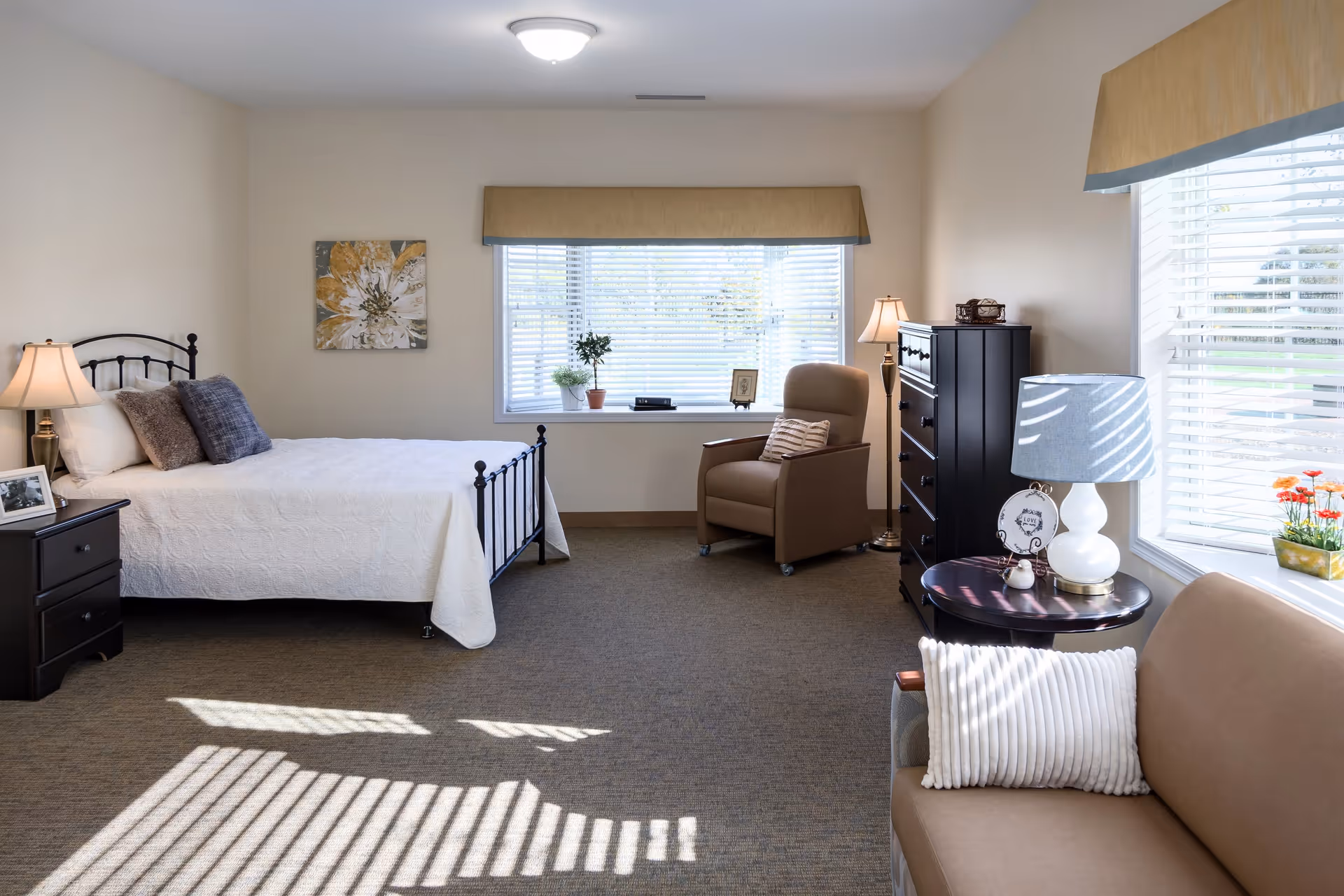 A bright and spacious bedroom in a senior living facility featuring a neatly made bed with white bedding and decorative pillows, a nightstand with a lamp and framed photo, a comfortable armchair near a large window with blinds, a dark wooden dresser, a round side table with a lamp, and a beige sofa with a white pillow. The room has beige walls and carpeted flooring.