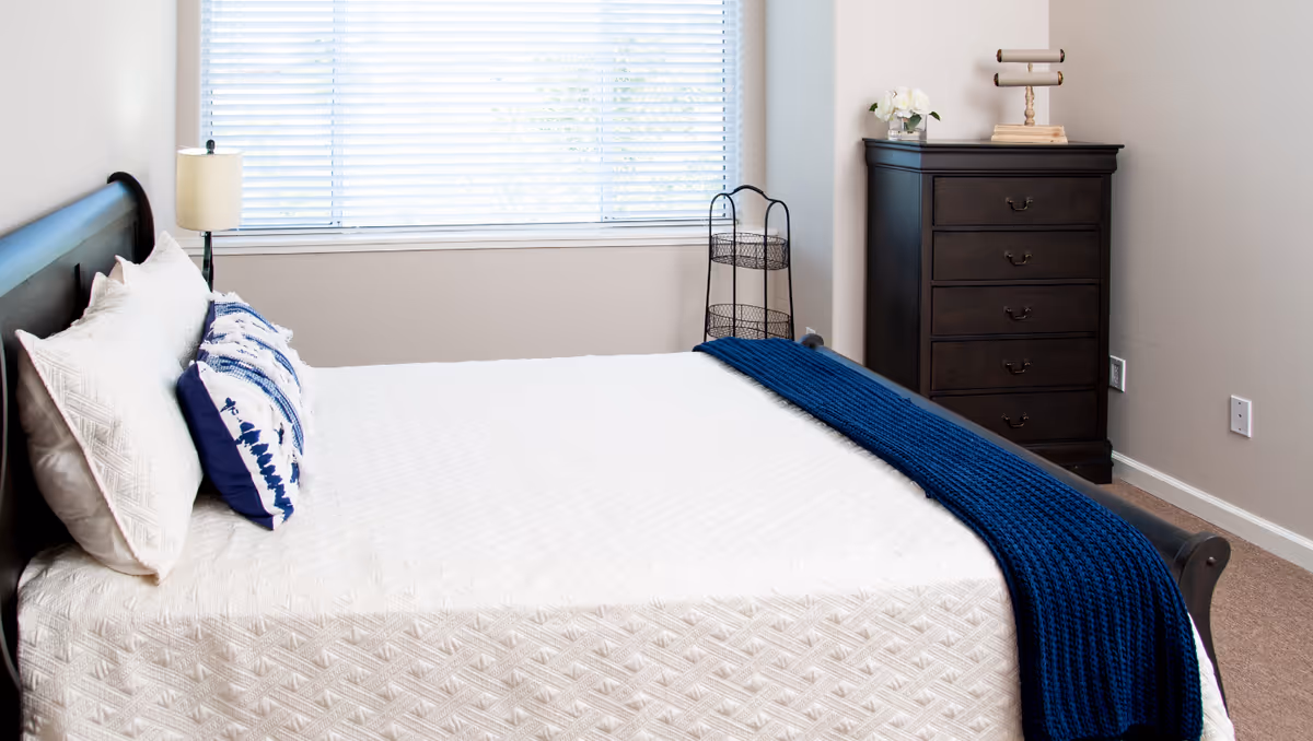 Bright bedroom with a made bed covered in a white quilt and blue throw, a bedside lamp, and a dark wood dresser beneath a window.