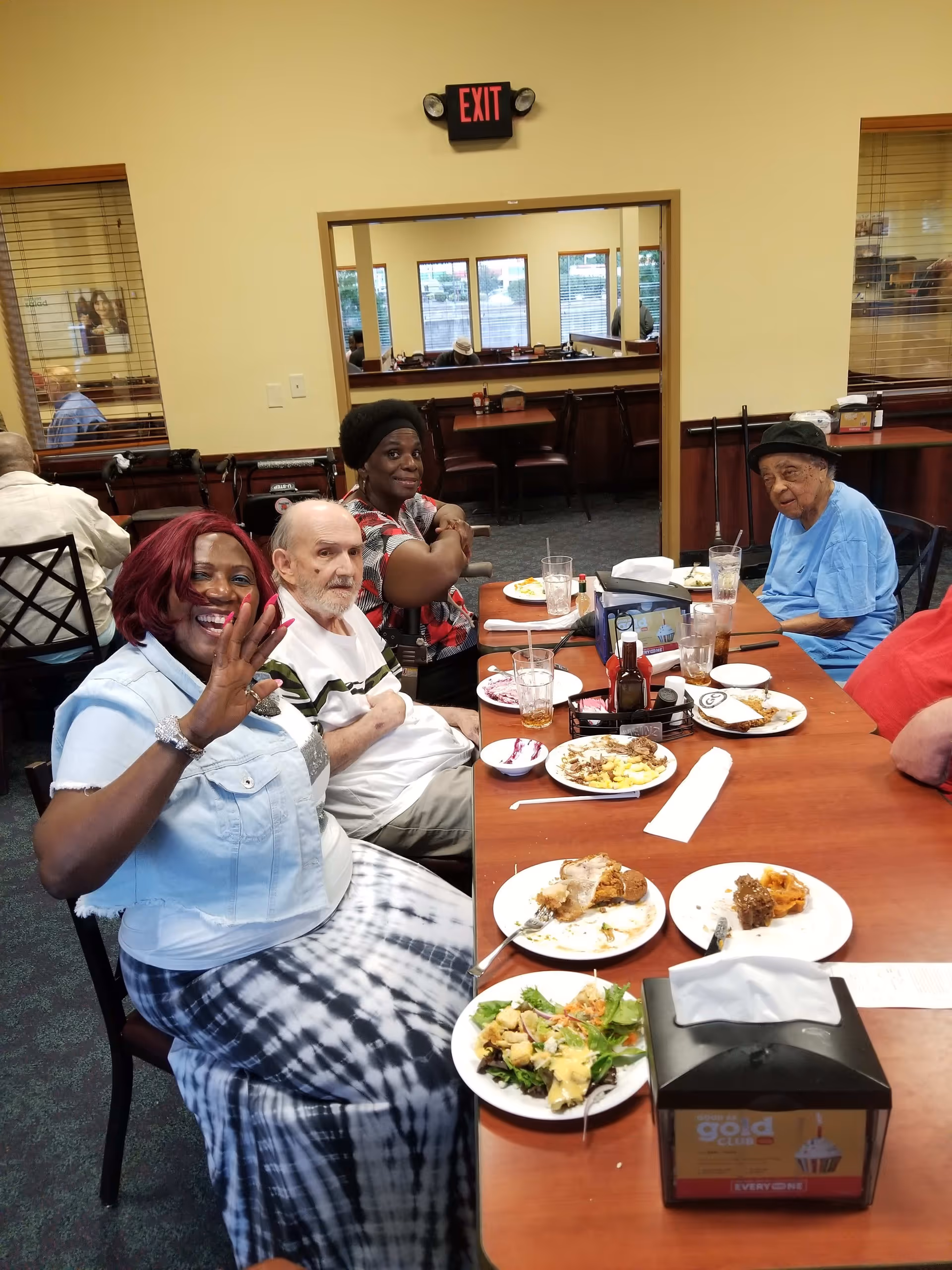 A group of elderly people sitting around a dining table in a senior living facility, enjoying a meal together. One woman in the foreground is smiling and waving at the camera. Plates with food, glasses, and condiments are on the table. The room has large windows and an exit sign above a doorway.