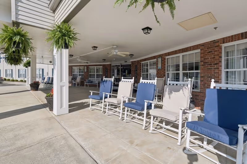 Covered outdoor patio area with white rocking chairs featuring blue and beige cushions lined up along a brick building. Hanging green ferns are suspended from the ceiling, and ceiling fans are installed above. The area is well-lit with natural sunlight.