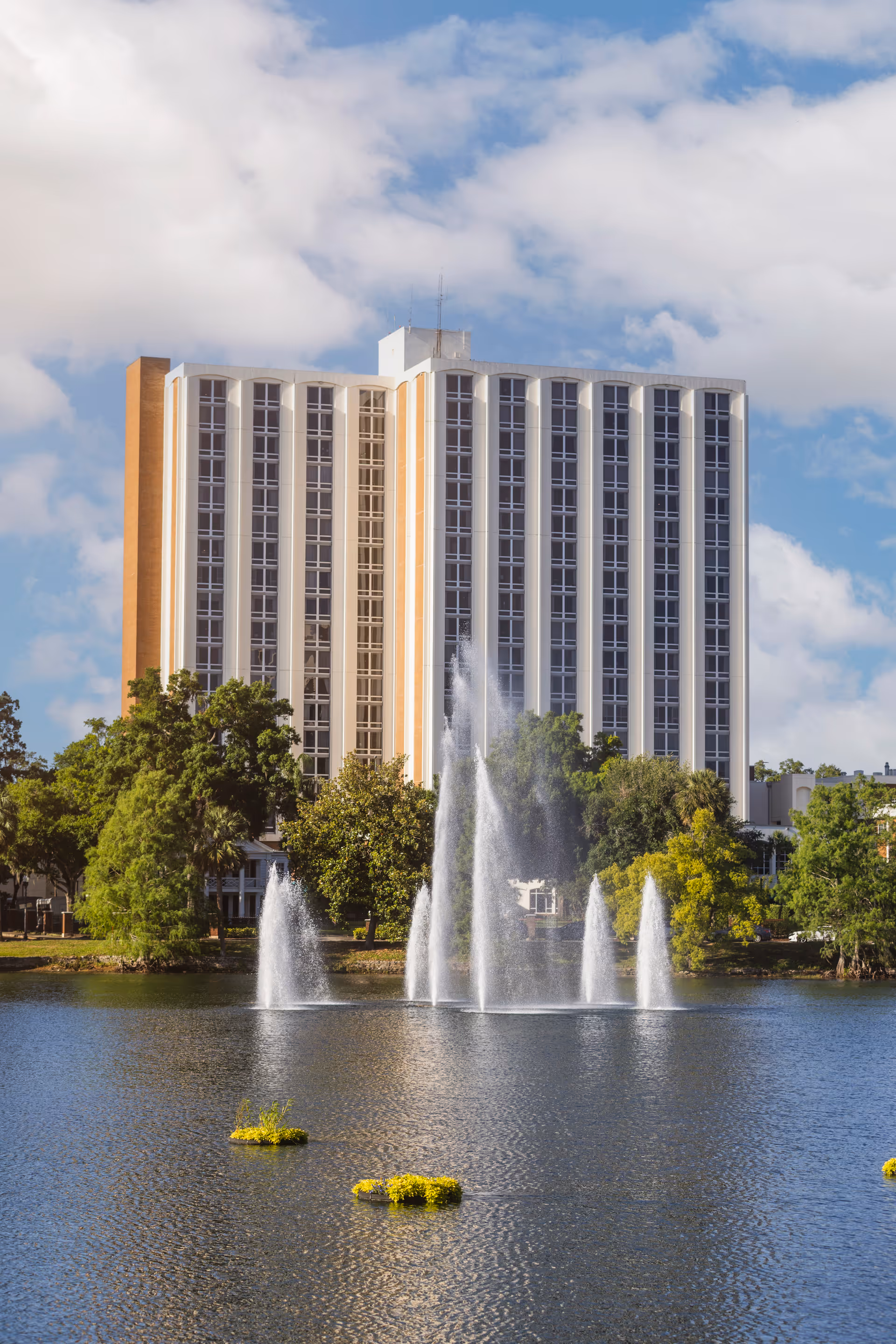 Tall multi-story building by a lake with several water fountains and surrounding trees under a partly cloudy sky.