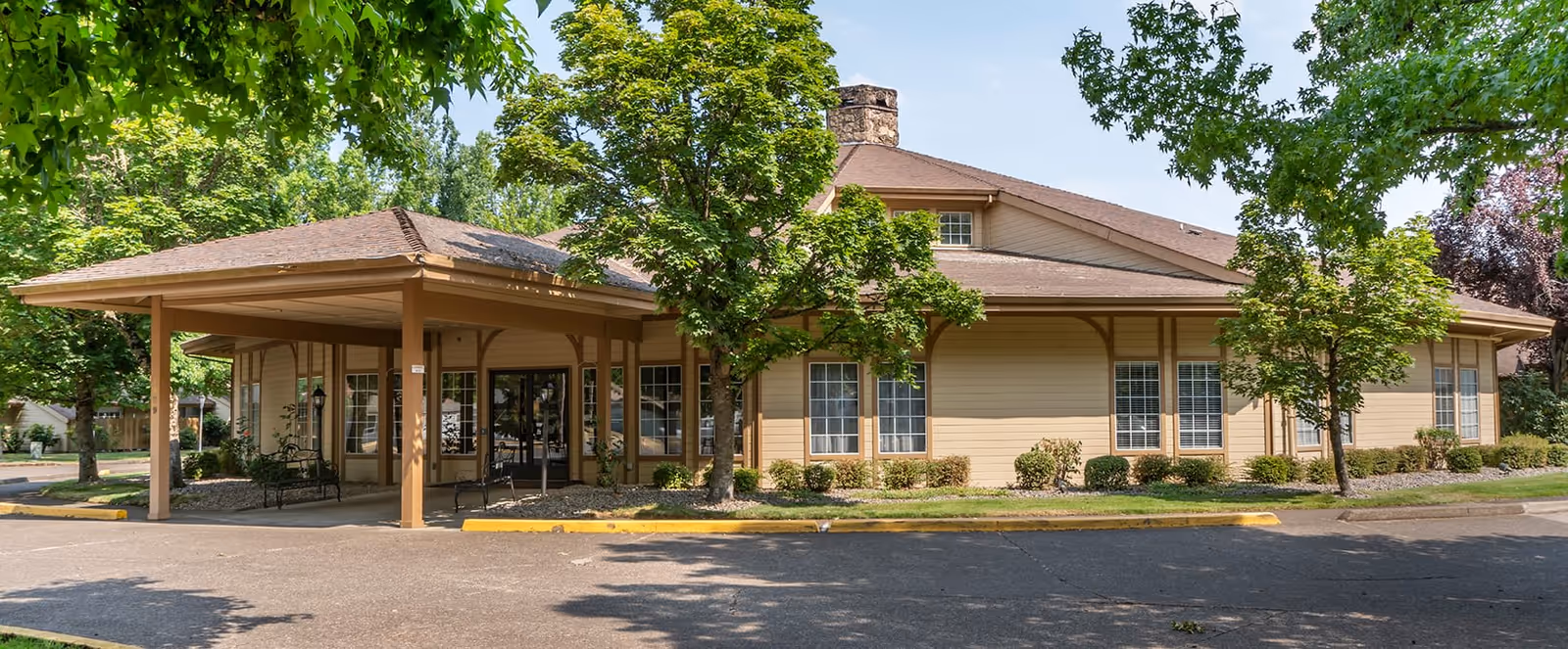 Exterior view of a single-story building with a covered entrance, surrounded by trees and shrubs under a clear blue sky.