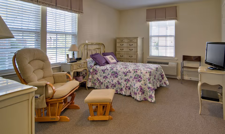 A cozy bedroom in a senior living facility featuring a single bed with a floral purple and white bedspread, a cushioned rocking chair with a matching footrest, a nightstand with a lamp and phone, a dresser, a TV on a small table, and two windows with blinds and valances.