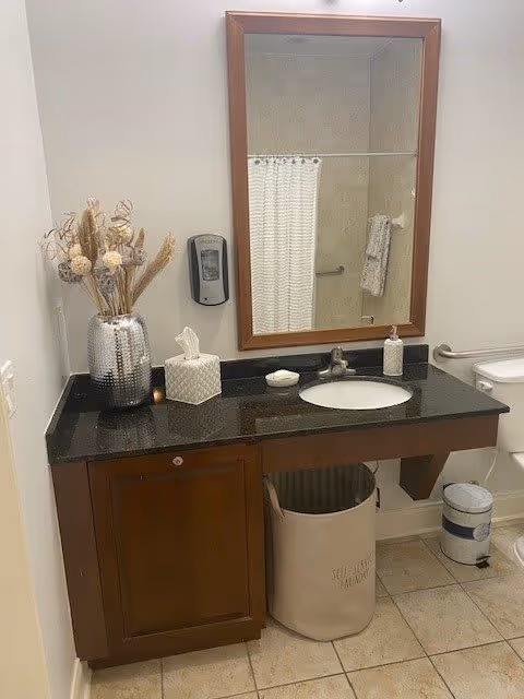 A bathroom vanity with a dark granite countertop, an oval sink, and a wooden cabinet below. On the countertop, there is a silver vase with dried flowers, a tissue box, a soap dish with soap, and a liquid soap dispenser. Above the vanity is a large rectangular mirror with a wooden frame. To the right, a toilet is partially visible with a grab bar on the wall next to it. The floor is tiled, and a beige laundry hamper is placed under the vanity.