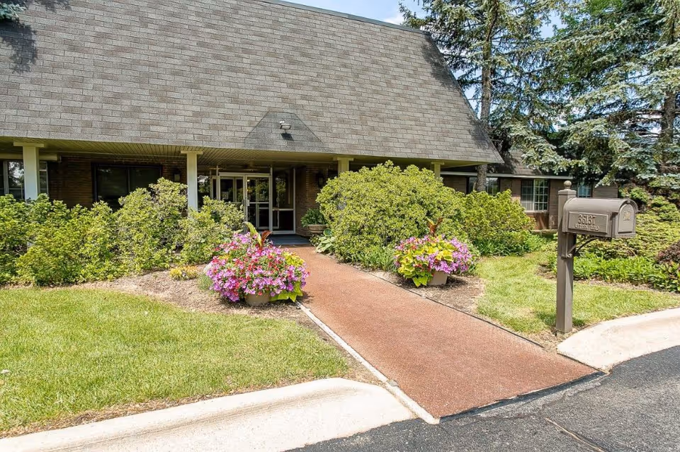 Entrance to a senior living facility with a sloped roof, surrounded by green bushes and colorful flower pots on either side of a paved walkway. A mailbox is visible on the right side near the curb.