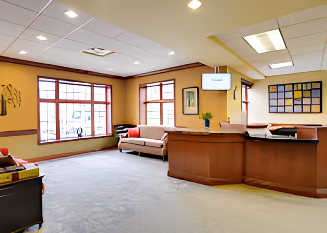 A spacious interior room with large windows letting in natural light, featuring a wooden reception desk with a small plant on it, beige sofas with red cushions, framed artwork on the walls, and a carpeted floor.